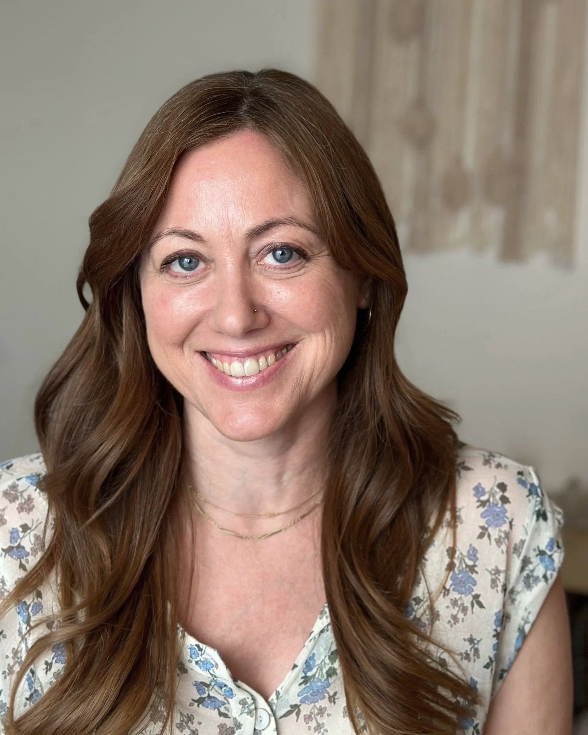 A woman with long red hair, blue eyes, and a nose piercing, smiling at the camera, wearing a floral blouse and layered necklaces.