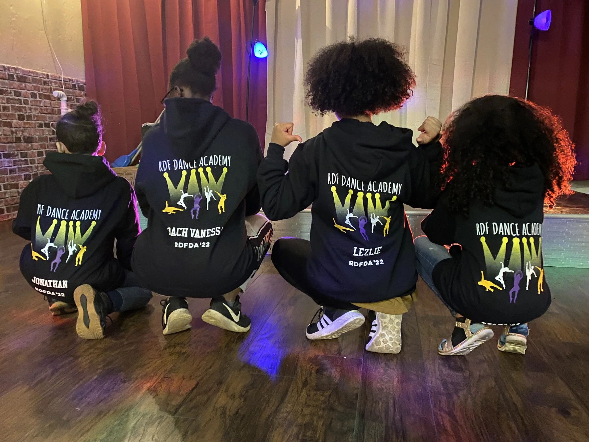Four children sitting on the floor with their backs facing the camera, wearing matching black hoodies with colorful dance silhouettes and text that reads 'RDF Dance Academy' and their names, inside a dance studio with brick wall and stage lights.