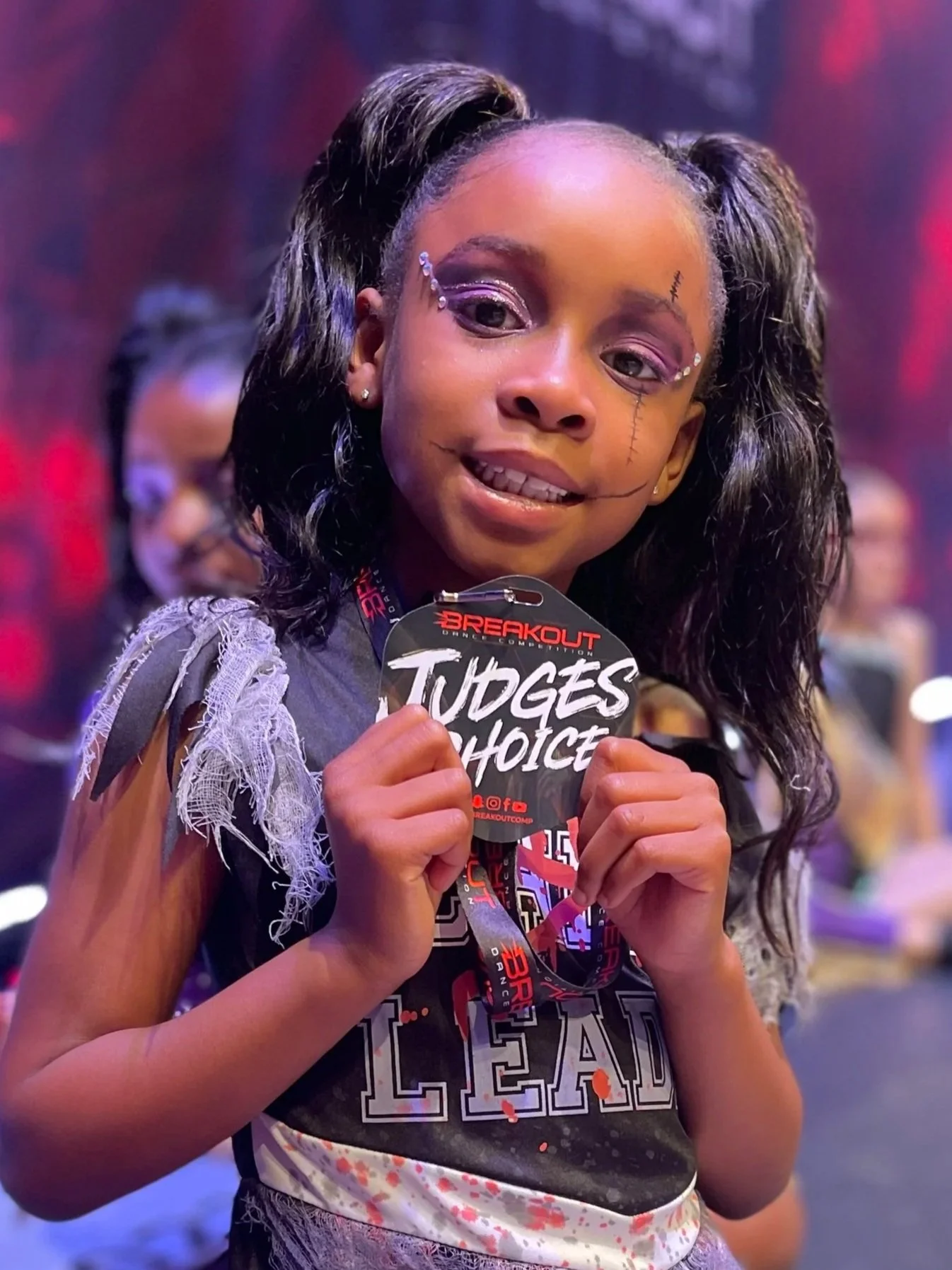 Young girl at a competitive event holding a medal with 'Judges Choice' on it, wearing makeup and costume with face paint and accessories, smiling with other children in the background.
