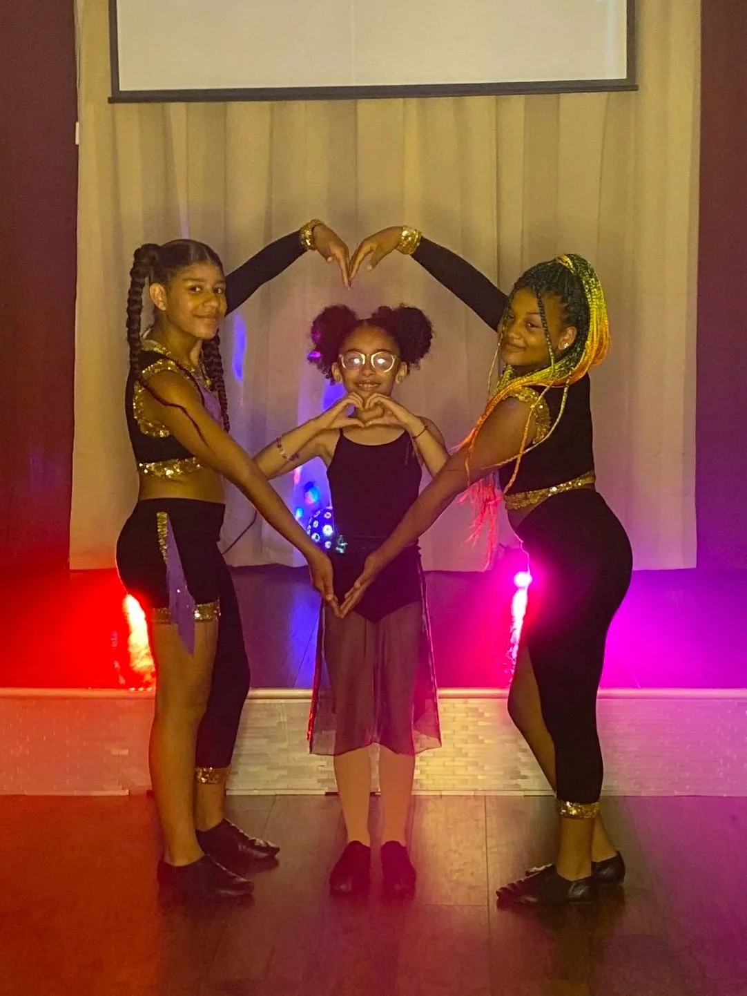 Three young girls dressed in dance costumes posing on a stage, making a heart shape with their hands and arms. Nova Dance Academy