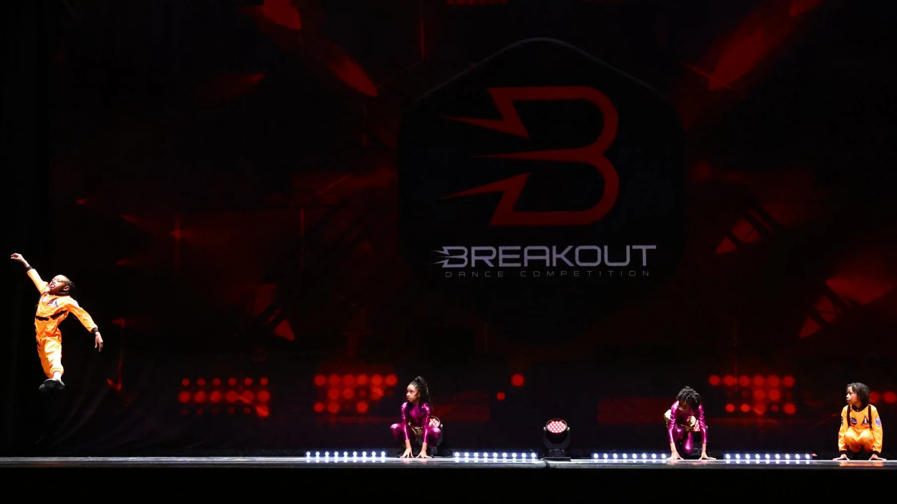 Nova Dance Academy's young dancers perform on stage during a break out dance competition, with a large screen in the background displaying the event logo.