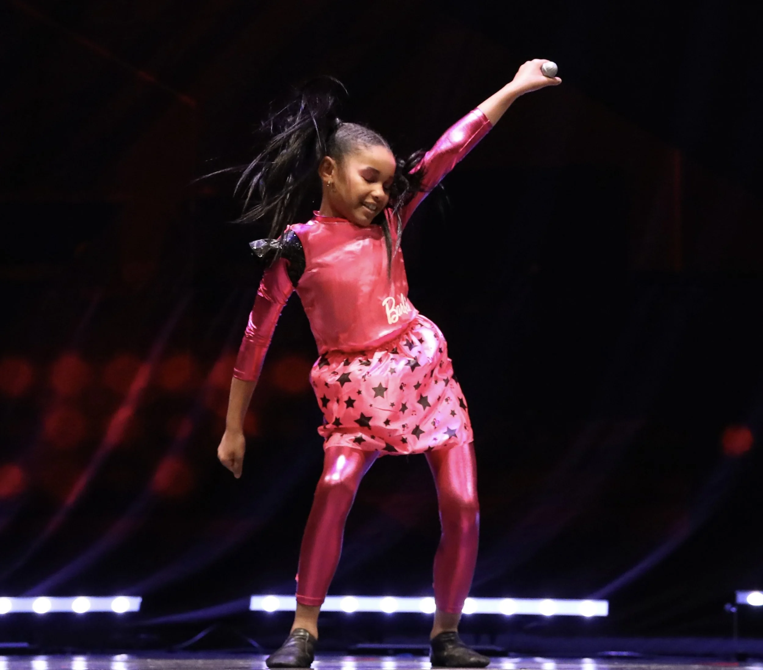 Young girl performing on stage, wearing pink and black costume with star patterns, holding a microphone with styled hair.