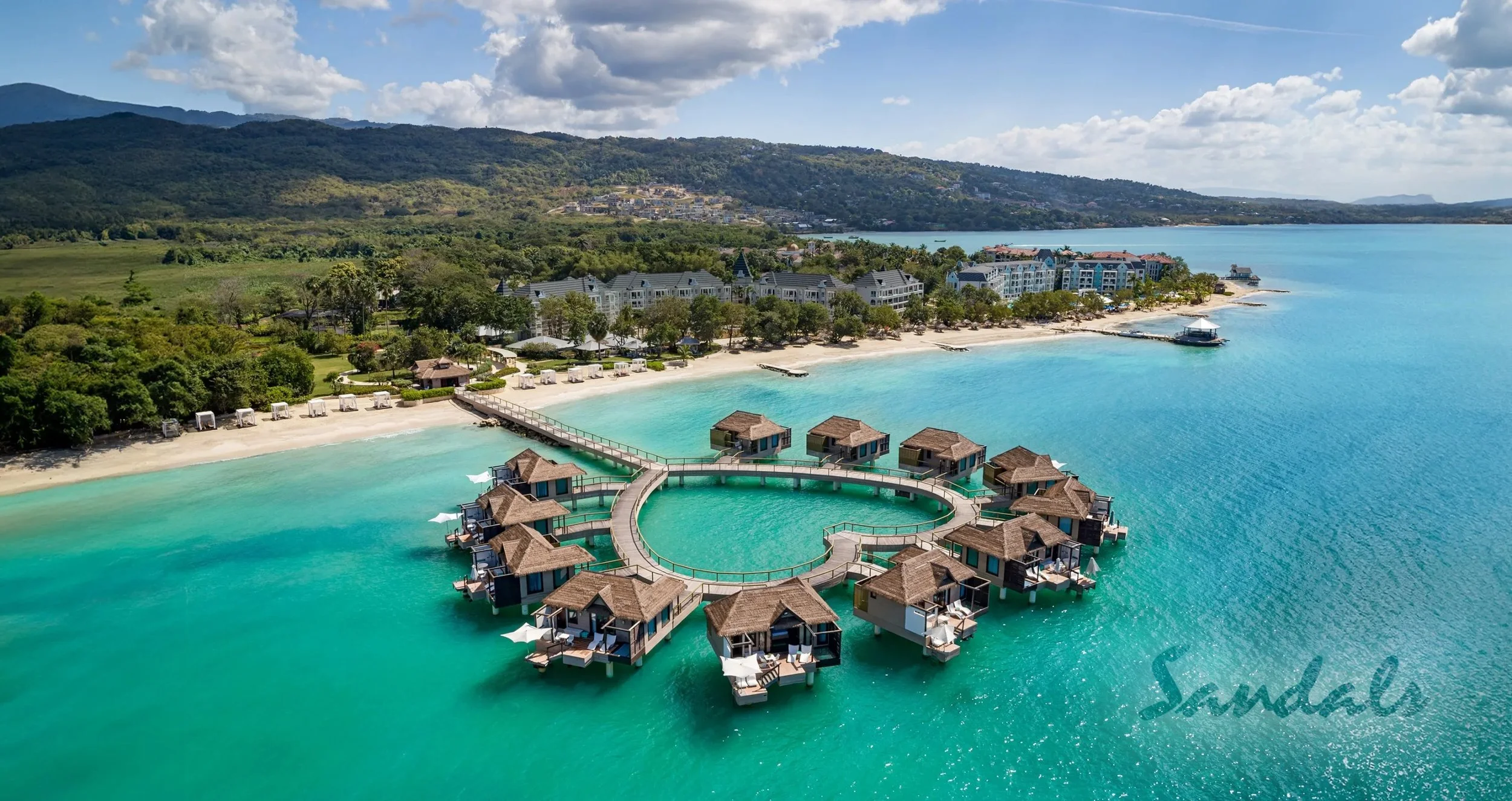 Overwater bungalows in a tropical beach resort with turquoise water, white sandy beach, lush greenery, and mountains in the background.