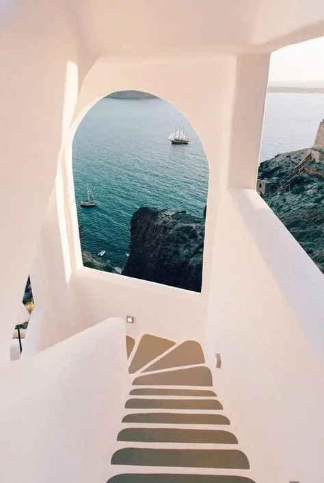 View of the sea with sailboats through an arched window in a white building, with curved stairs leading down.