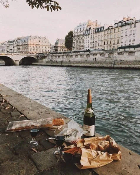 Picnic setup with wine, pizza, and a bottle along a riverbank in Paris, France, with buildings and a bridge in the background.