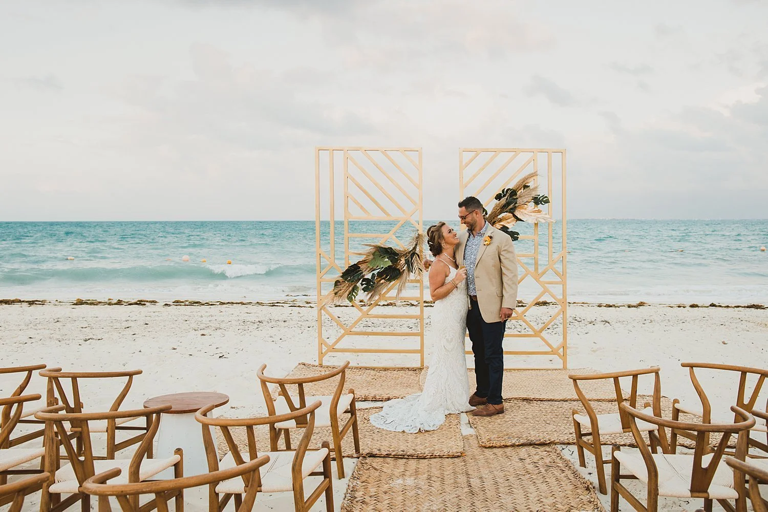 A bride and groom stand together on a beach wedding altar made of geometric wooden panels decorated with dried plants and greenery, with ocean in the background and empty chairs arranged for guests.