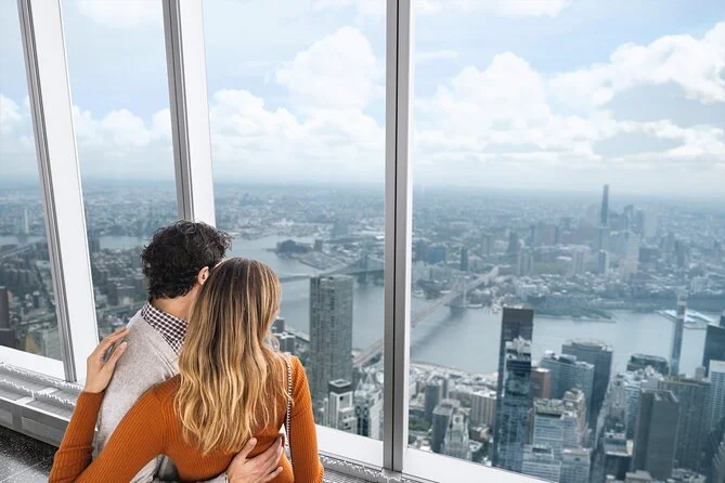 A couple looking out from a high observation deck at a city skyline, including a river and tall buildings, in daytime.