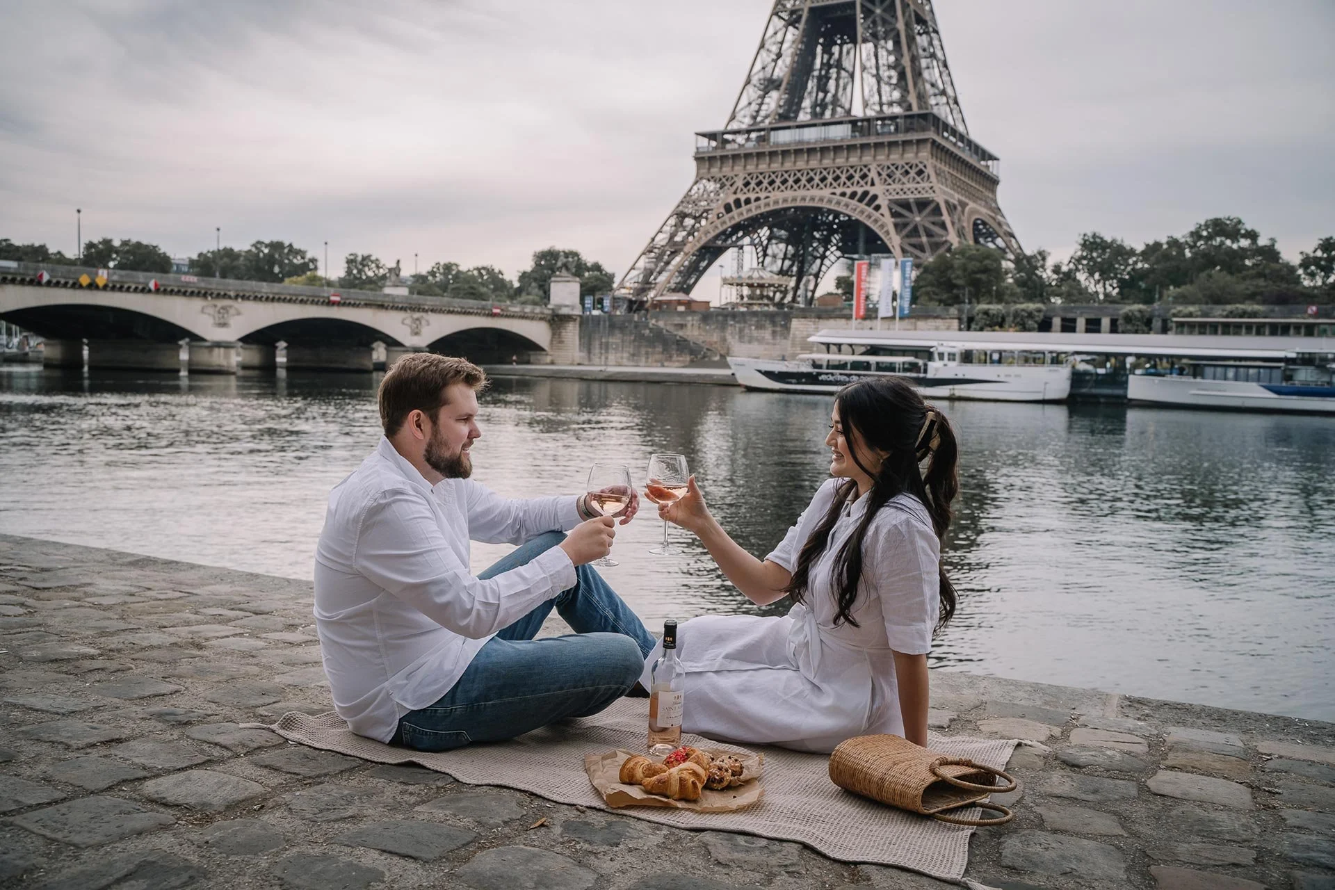 A couple enjoying a picnic on the Seine river bank with the Eiffel Tower in the background, toasting with glasses of rosé wine.