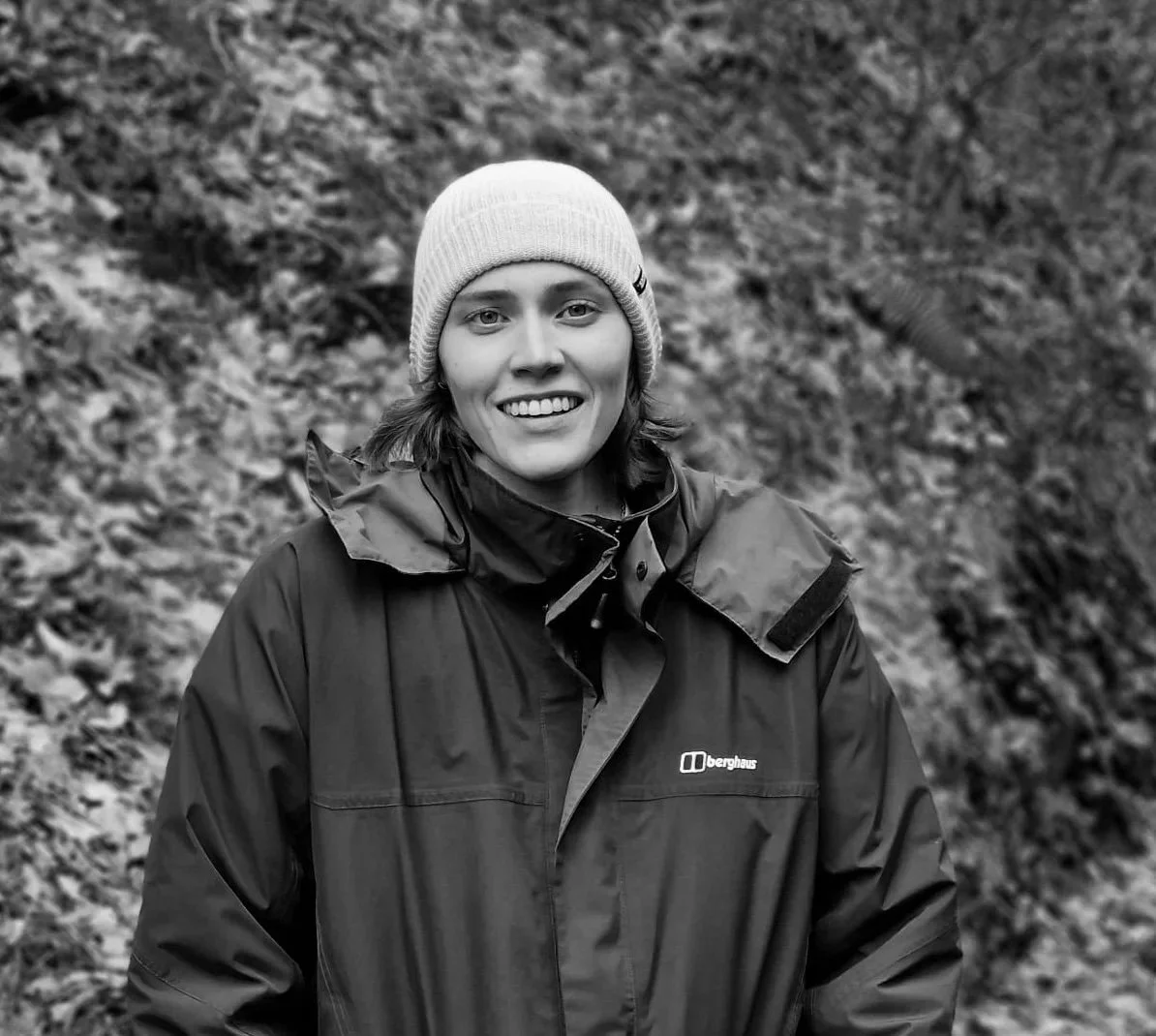 A smiling woman wearing a beanie and a Berghaus jacket outdoors with a background of fallen leaves.