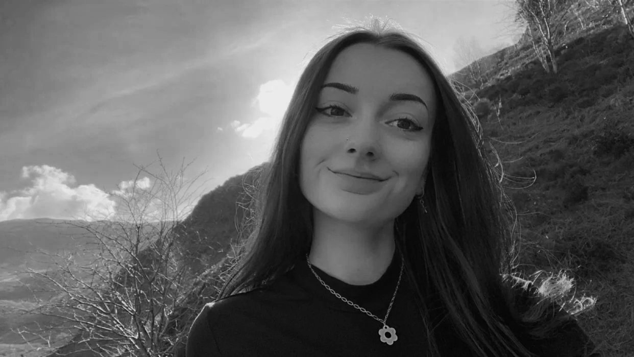 A black and white photo of a young woman with long dark hair smiling outdoors, mountains and cloudy sky in the background.