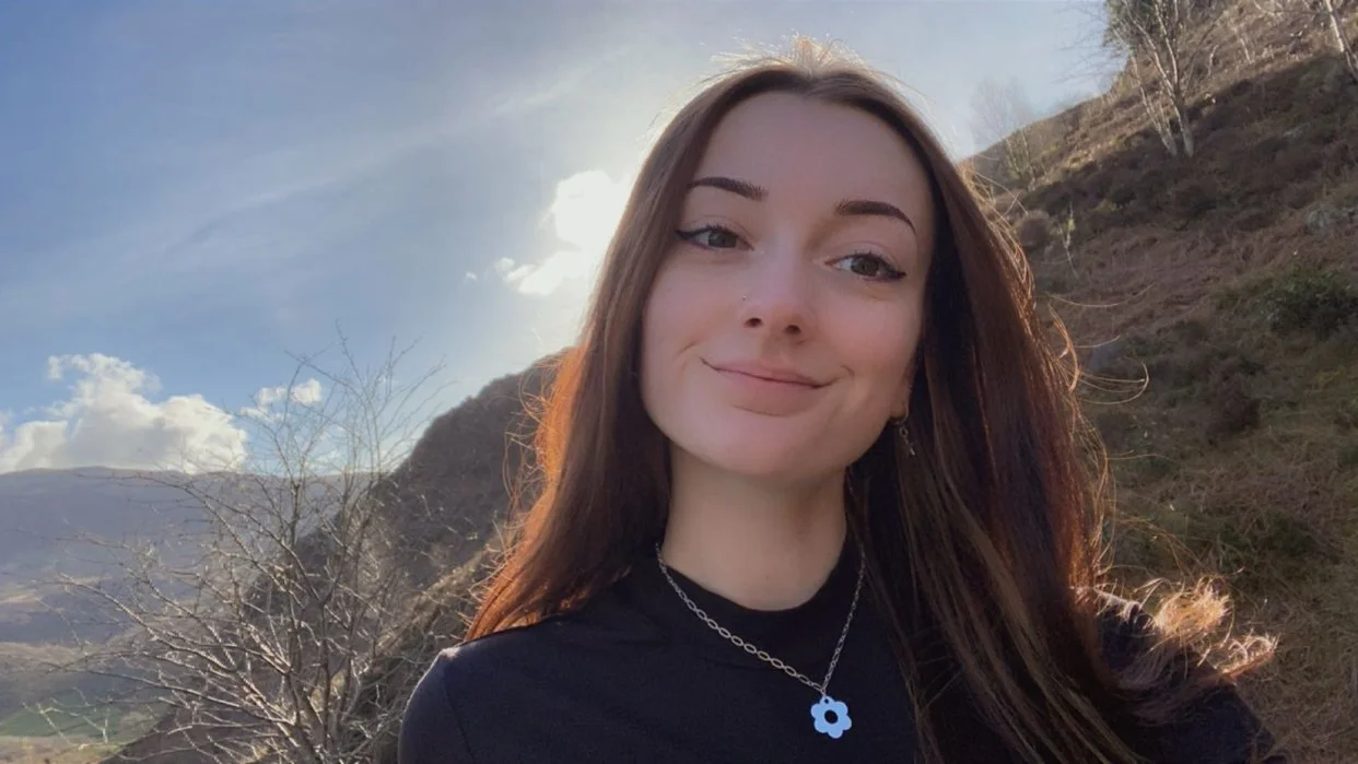 A woman with long brown hair smiling outdoors with hills and a partly cloudy sky in the background.