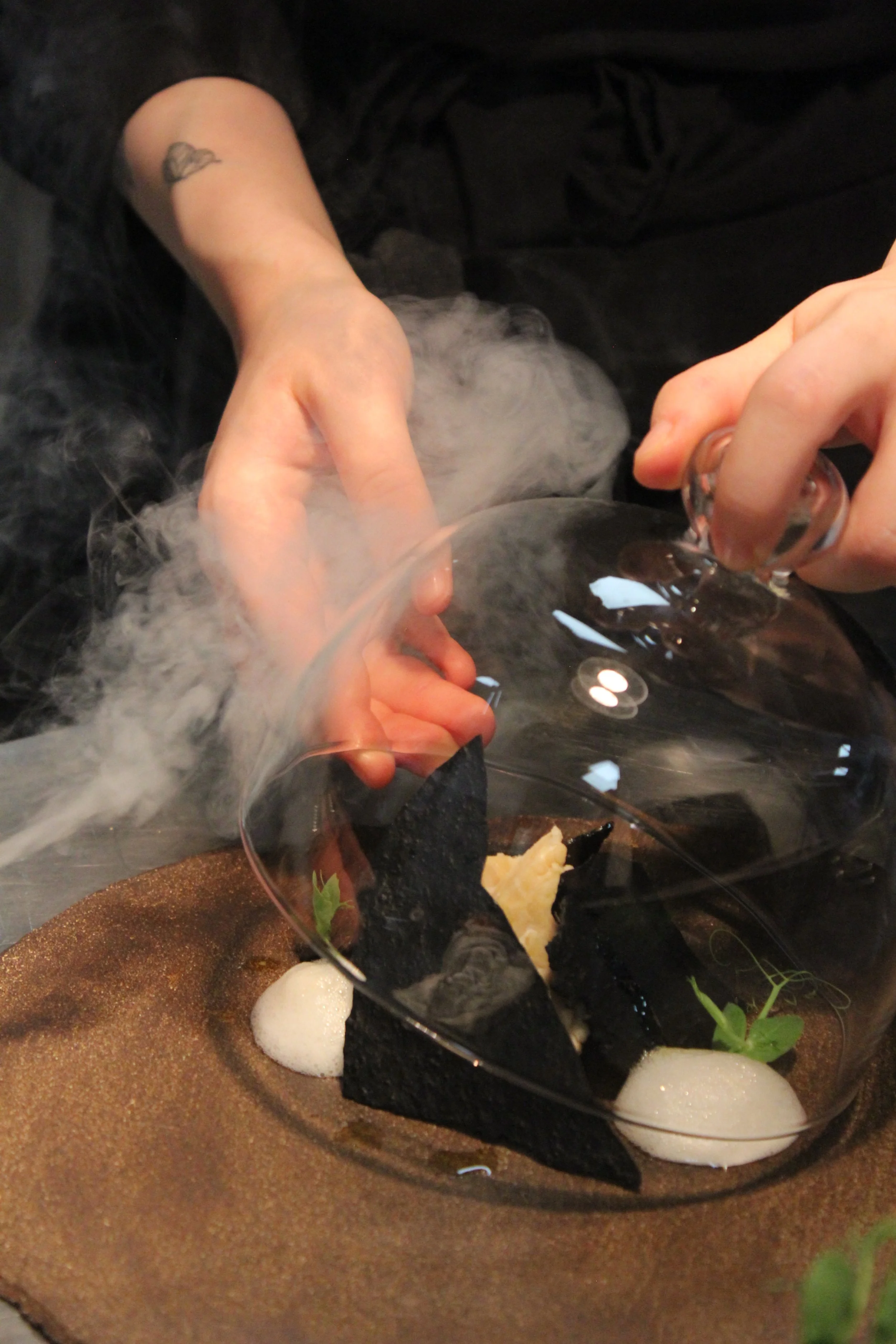 Close-up of hands lifting a glass cloche to reveal a smoking gourmet dish, symbolizing the theatrical and sensory nature of luxury fine dining.