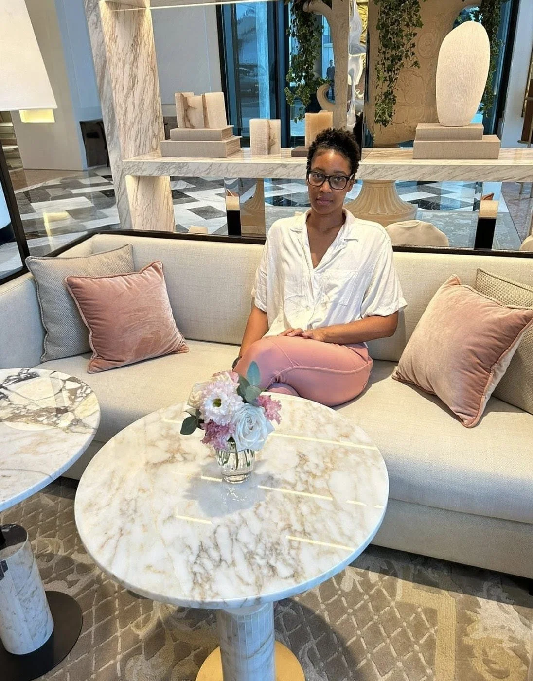 A professional portrait of a melanated woman in a white shirt and pink trousers seated on a beige sofa in a luxury hotel lobby. She sits behind a marble coffee table with flowers, framed by elegant shelving and decor.