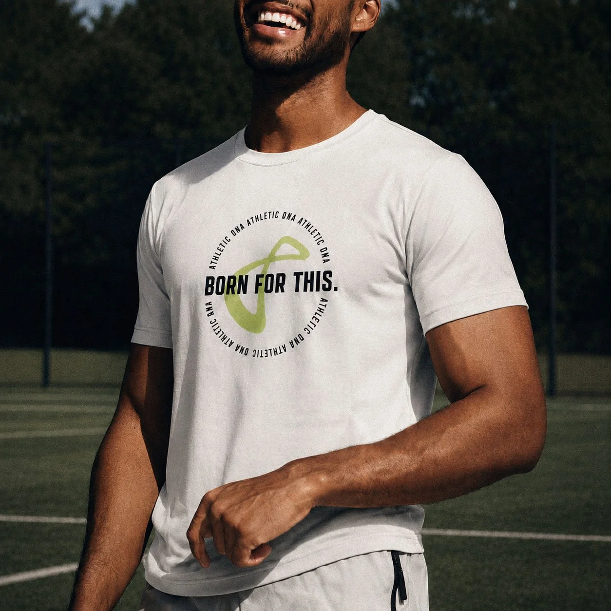 A man wearing a white athletic T-shirt with the phrase 'BORN FOR THIS' and a circular design, standing on a sports field during daylight, smiling.