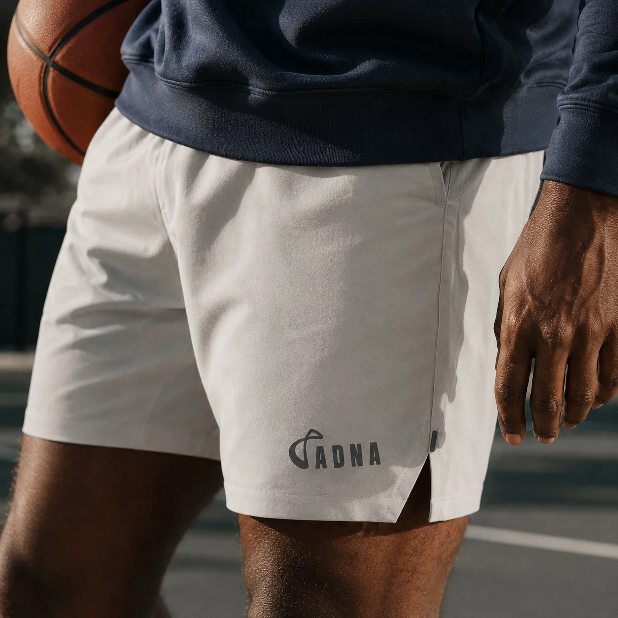 Close-up of a person wearing white athletic shorts with a logo that reads 'ADNA', holding a basketball in their left hand, standing outdoors on a basketball court.