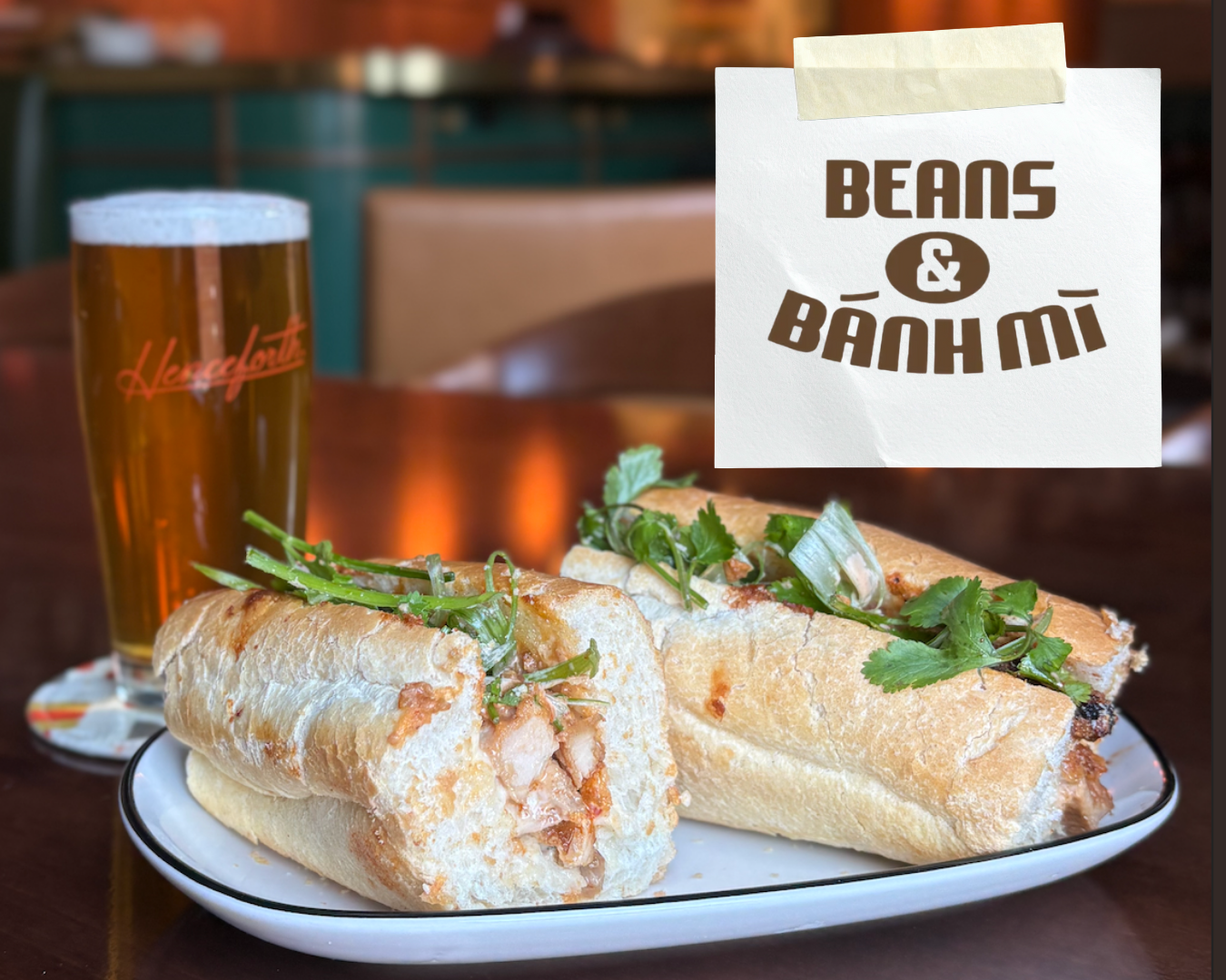 A plate with two banh mi sandwiches garnished with cilantro and green onions, a glass of beer, and a paper sign with the text 'Beans & Banh Mi' taped to the top right corner of the image.