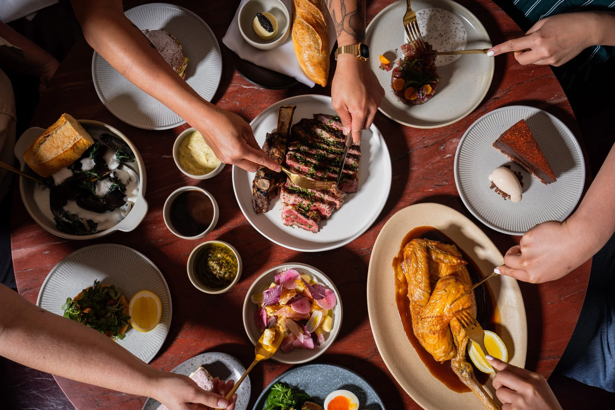People sharing a variety of dishes including steak, chicken, salad, and desserts around a wooden table.