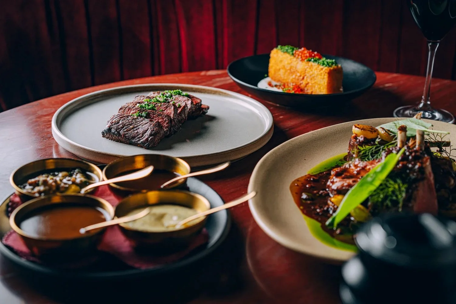 A variety of gourmet dishes on a wooden table, including a plate of grilled meat, a slice of corn casserole, a roast with greens and sauce, and three small bowls of sauces and accompaniments.