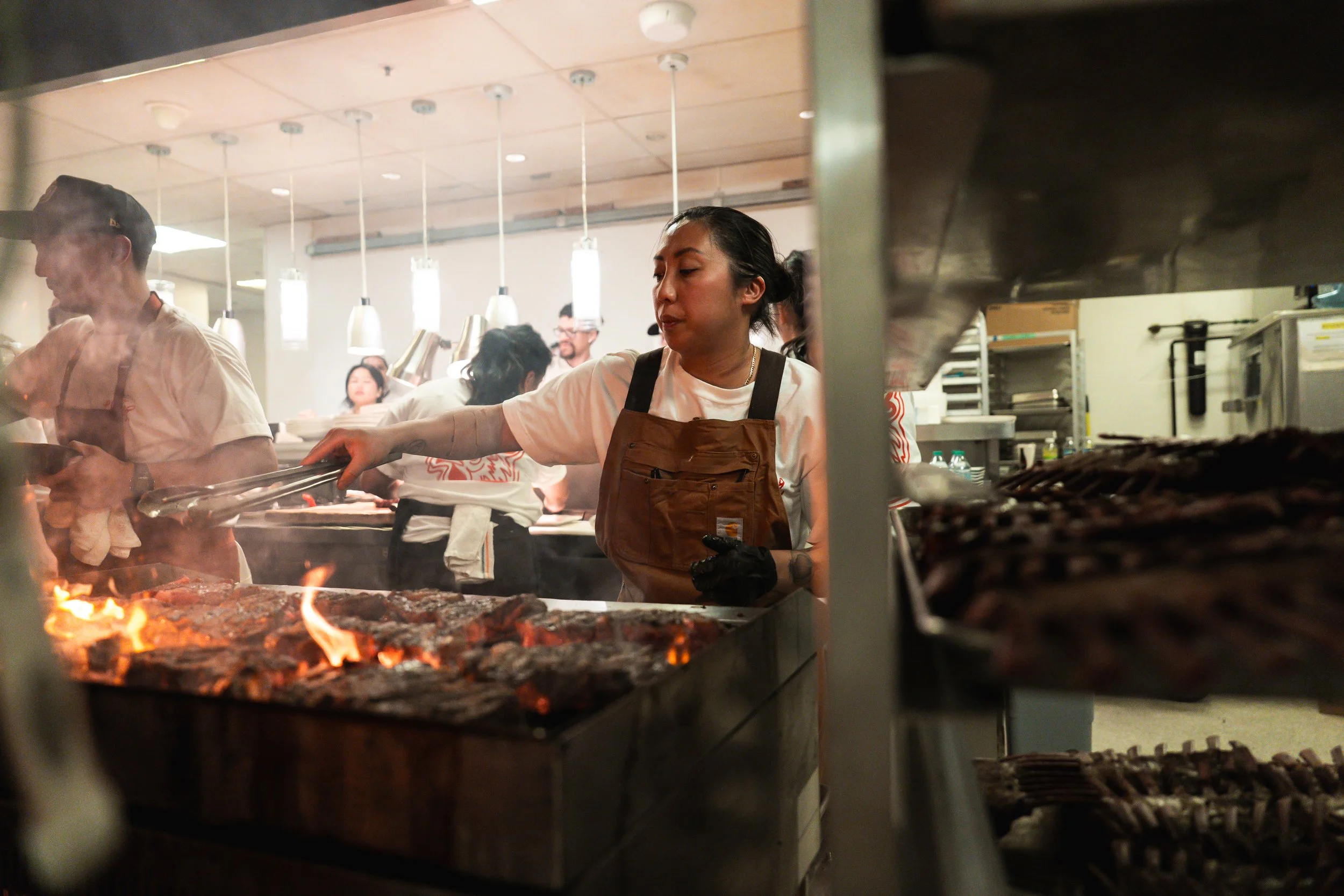 Tara Monsod wearing a brown apron cooks meat over an open flame grill in a busy kitchen.