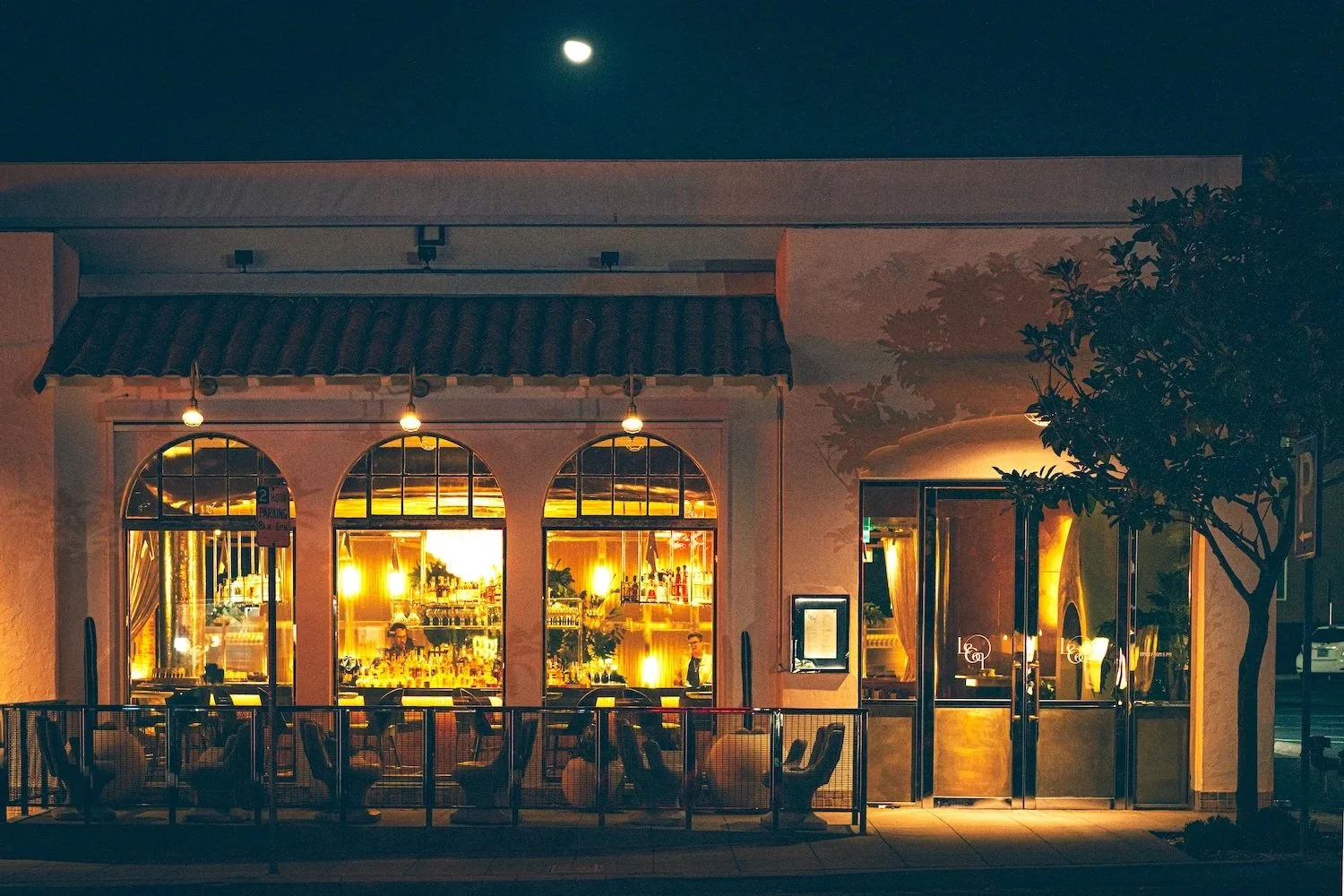 Night view of Le Coq restaurant in La Jolla with large arched windows, outdoor seating, and a tree beside the entrance; moon visible in the sky.