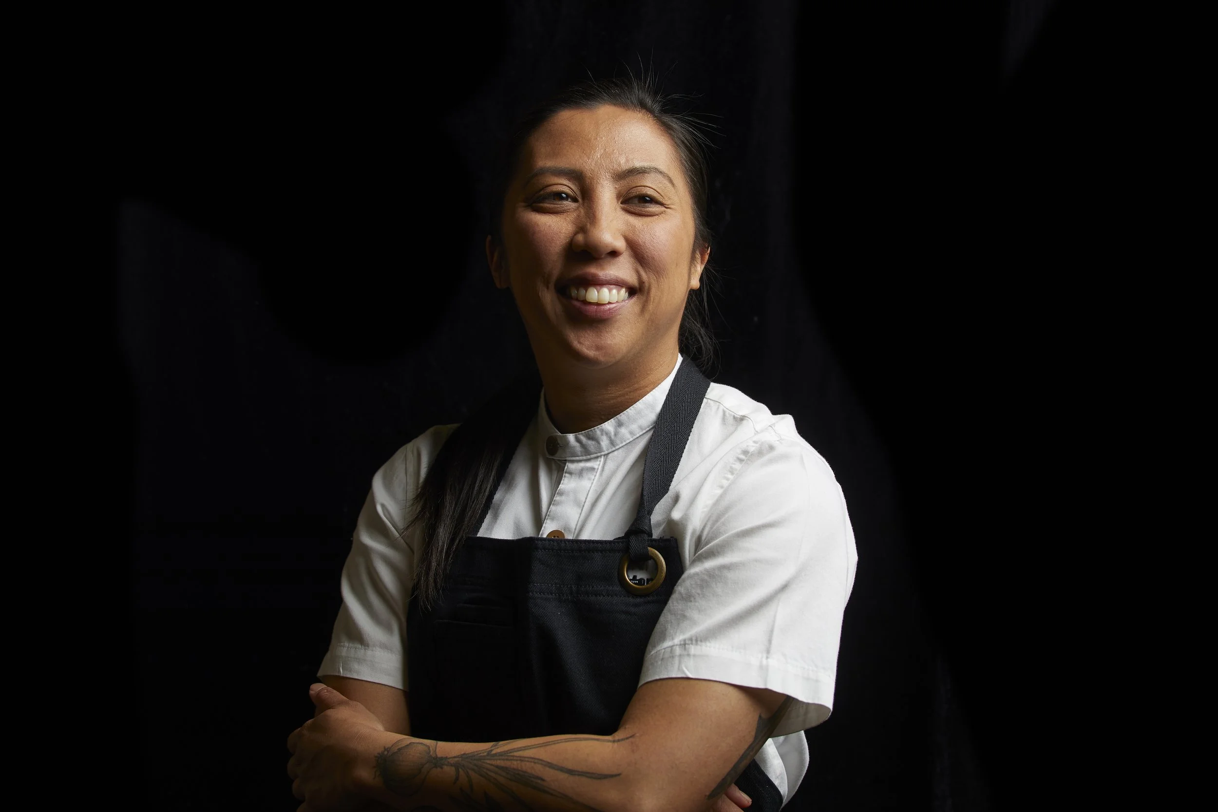 Chef Tara Monsod wearing a white chef's coat and black apron, smiling with arms crossed, against a black background.