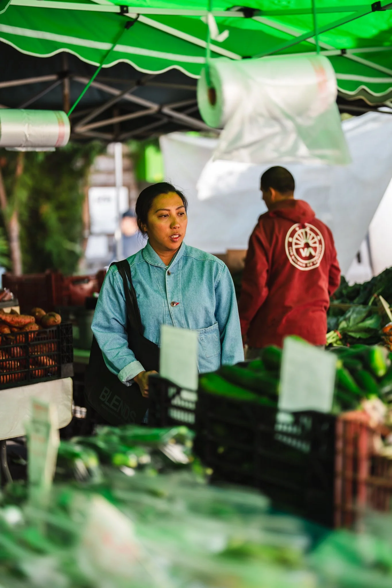 Tara Monsod shopping at a farmer's market in Little Italy San Diego with fresh vegetables on display, under a green canopy.