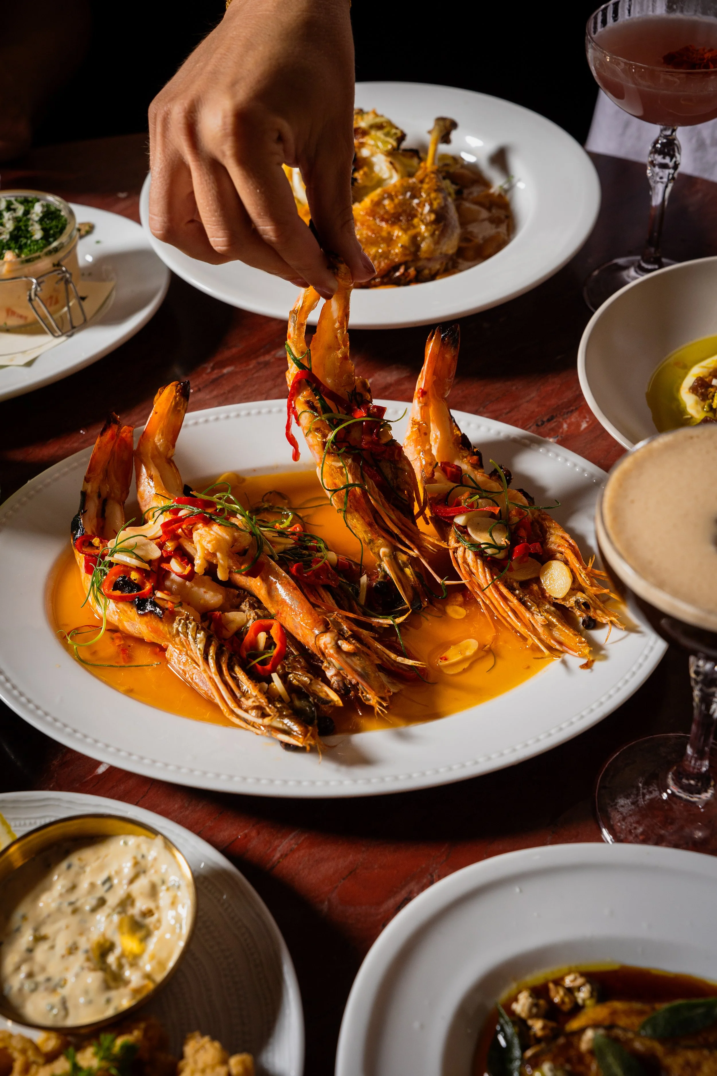 A plate of large cooked prawns garnished with sliced red chili and herbs, served in a spicy sauce, with a person’s hand picking up one of the prawns. Surrounding dishes include various appetizers, dips, and drinks on a wooden table.