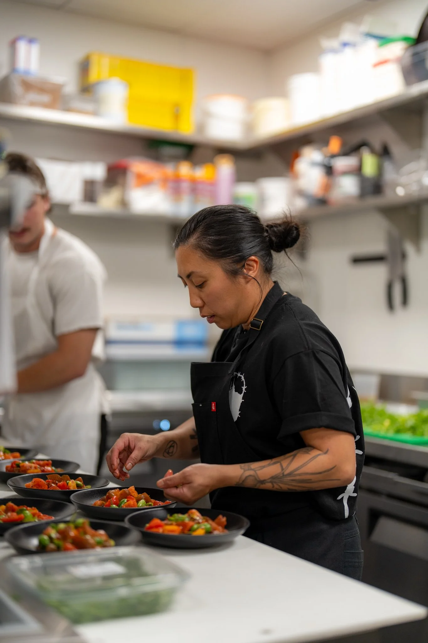 Chef Tara Monsod in a black apron preparing multiple black plates of food in a kitchen.