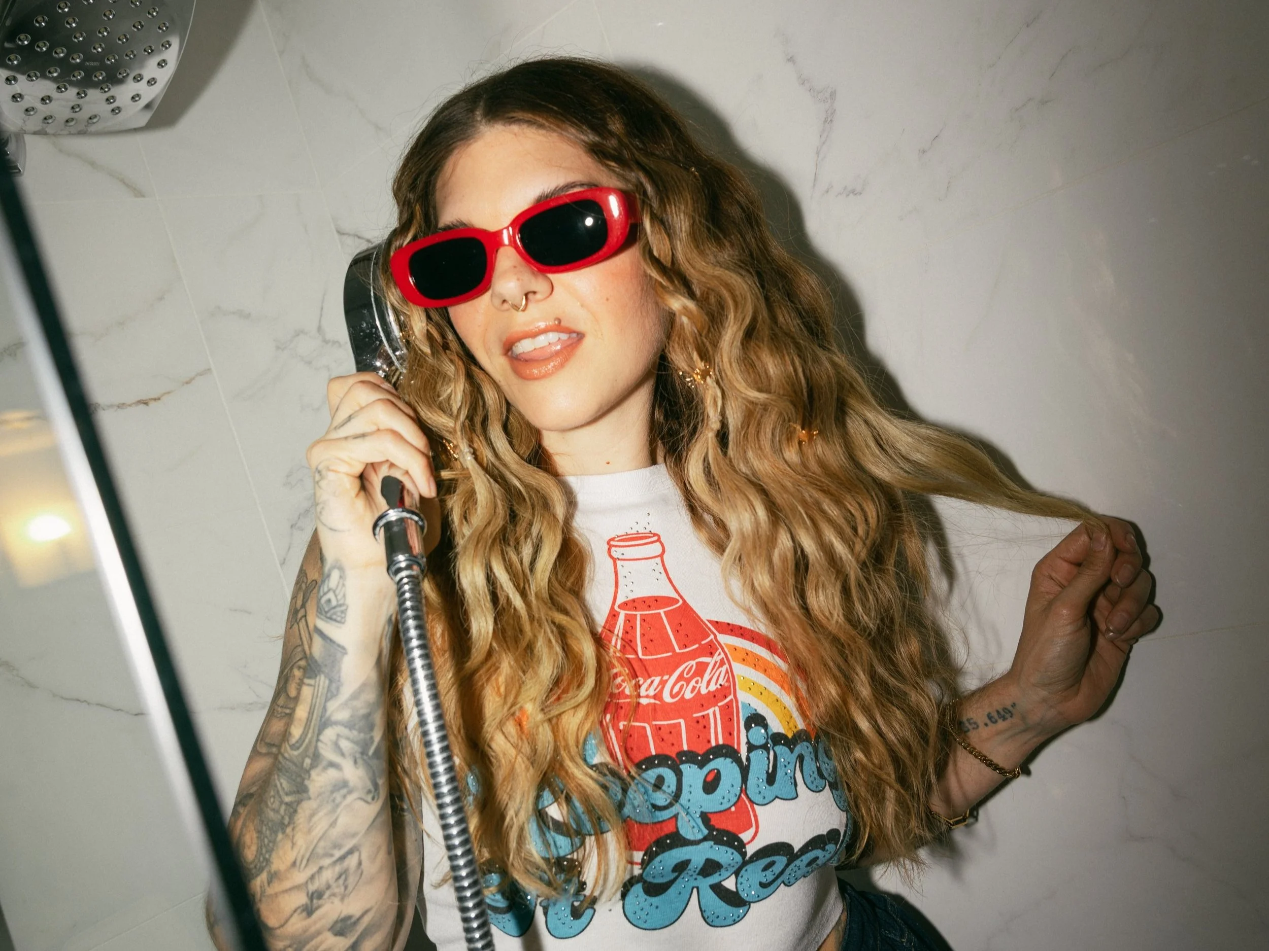 A young woman with long curly hair, wearing red sunglasses, a white T-shirt with Coca-Cola graphic and dark high-waisted pants, talking on a payphone in a bathroom.