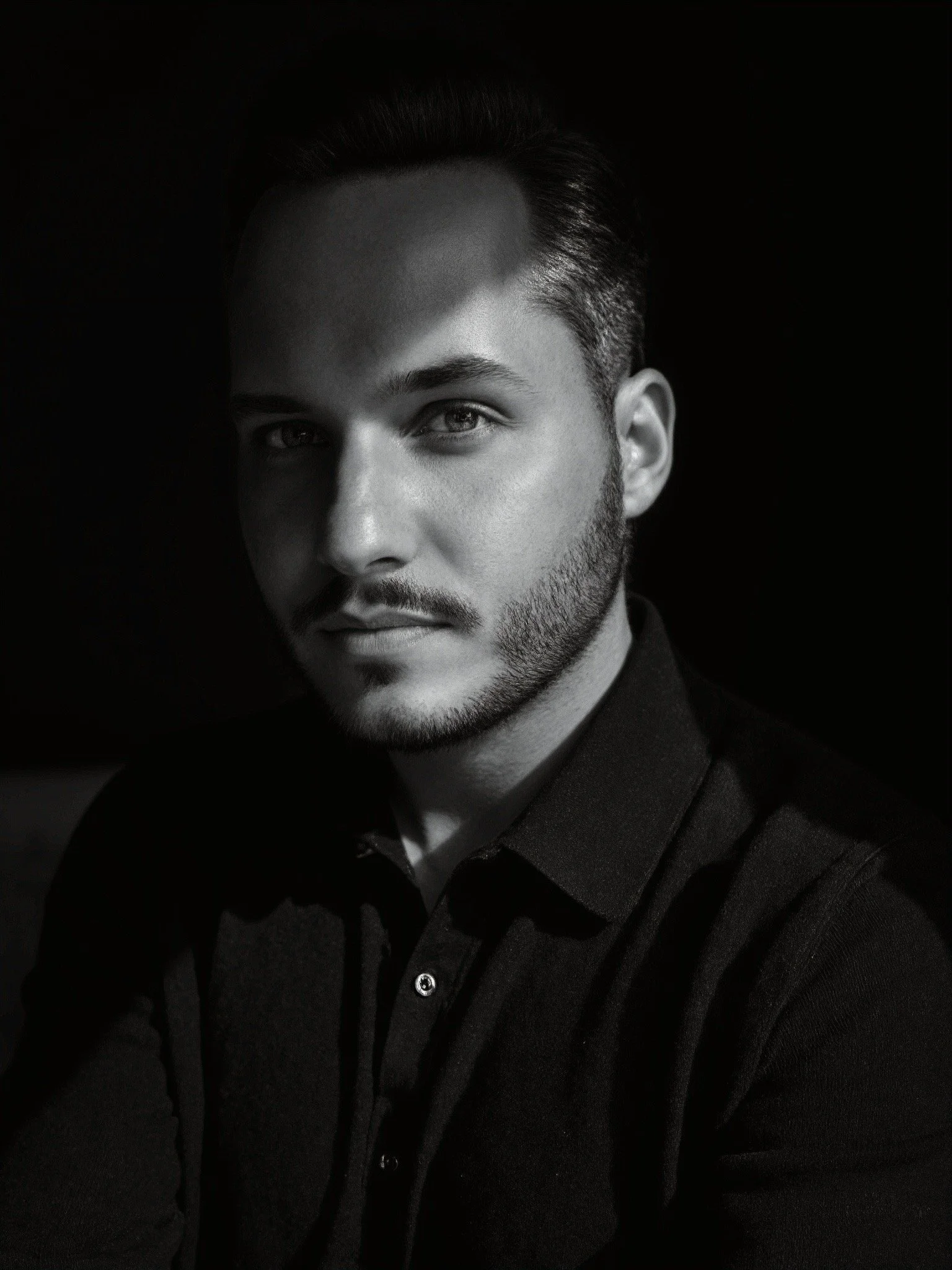 Black and white portrait of a young man with styled hair and facial hair, wearing a dark shirt, looking slightly to the side, against a dark background.