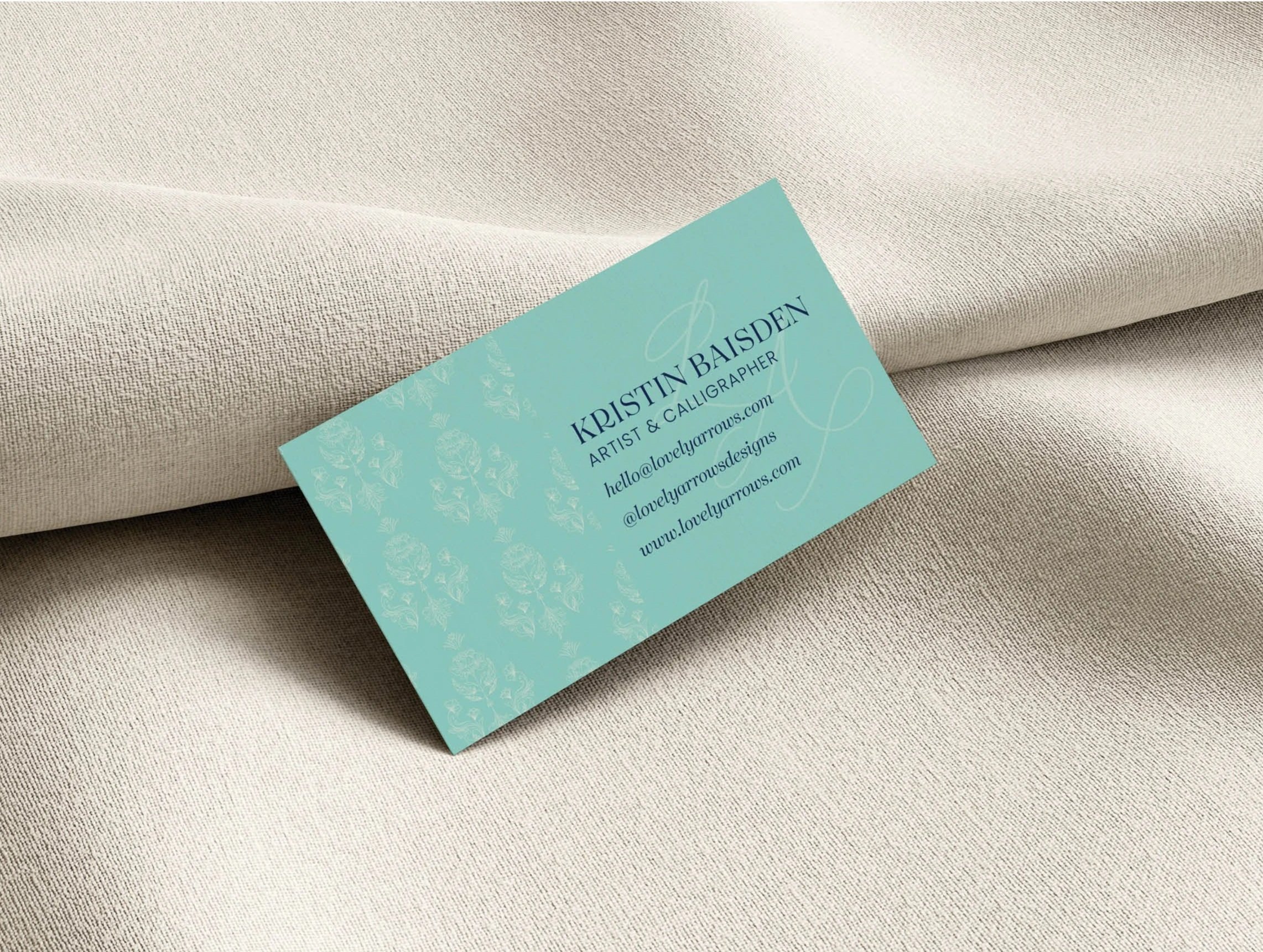A business card for Kristin Baisden, an artist and calligrapher, rests on a beige fabric surface.