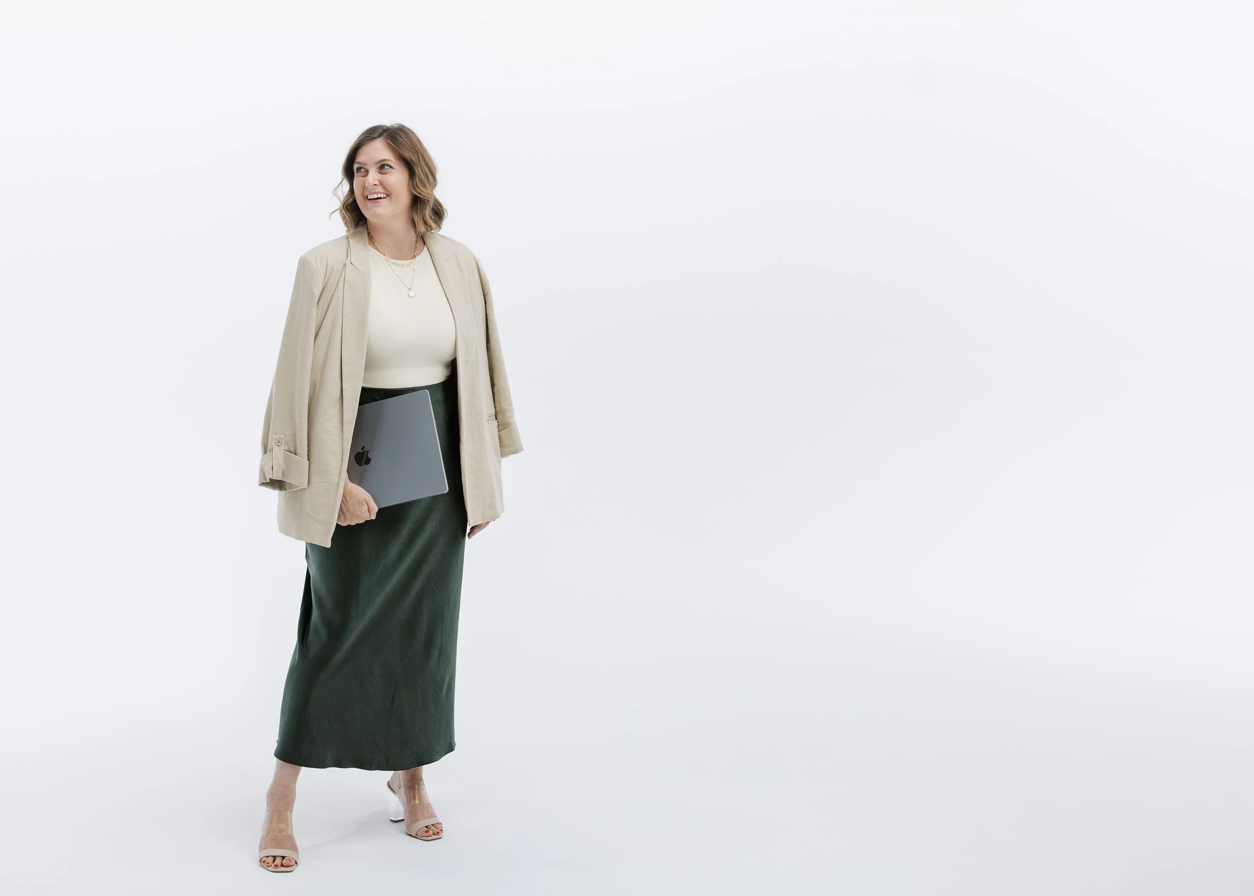 Jackie Burtch in a beige blazer and dark green maxi skirt holding a silver laptop stands against a plain white background, smiling and looking to her left.
