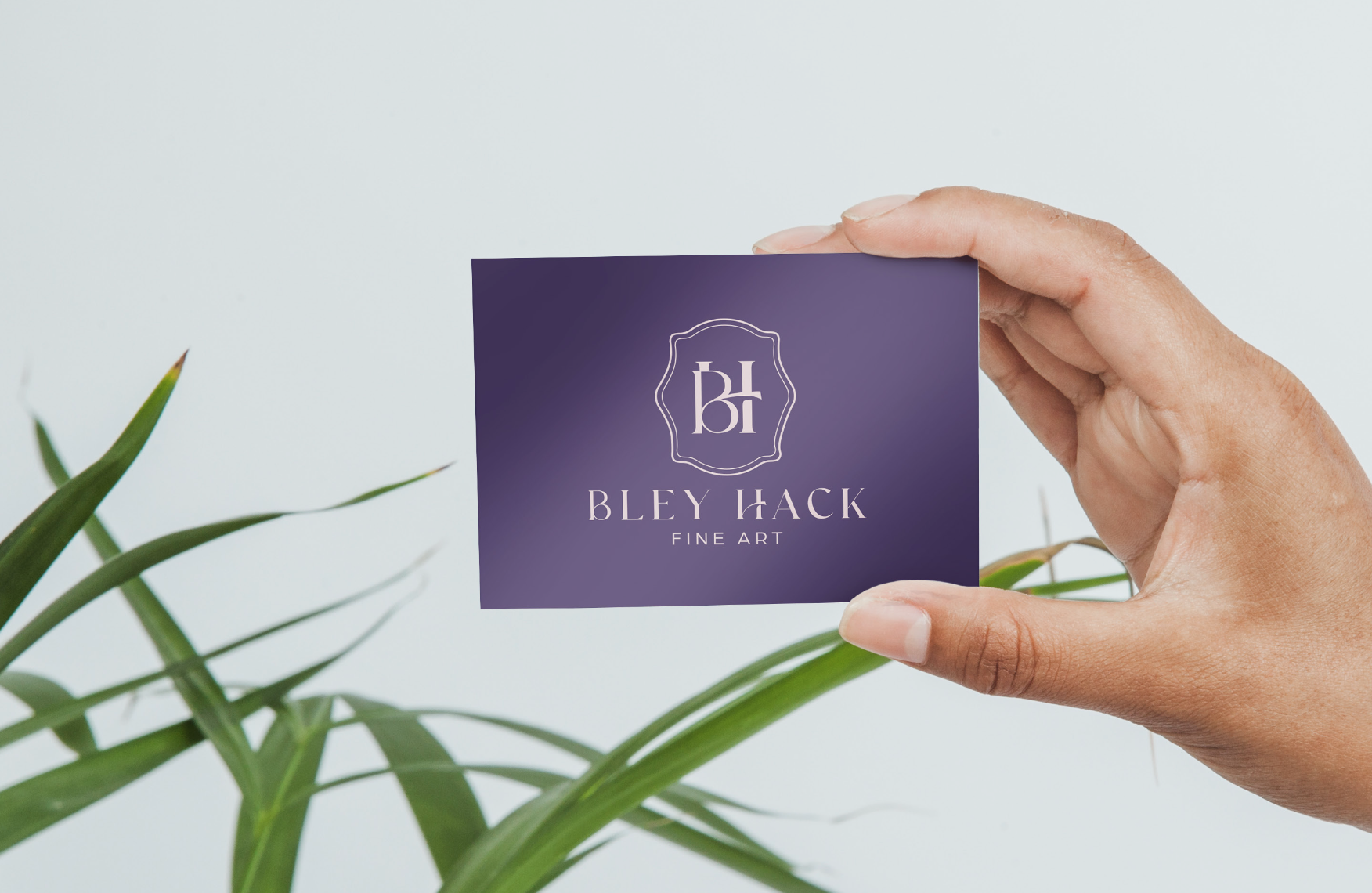 A hand holding a purple business card with the text 'Bley Hack Fine Art' and a logo, with green plant leaves in the foreground and a white background.