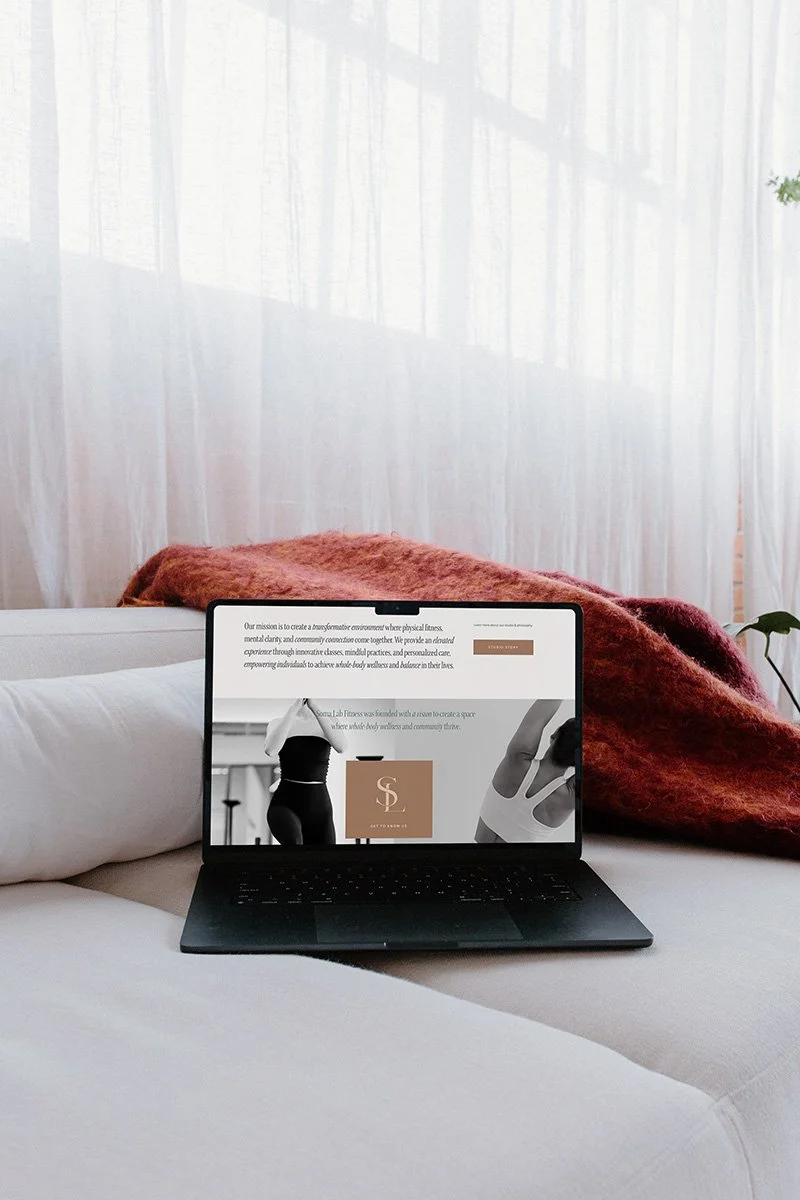 A black laptop on a white sofa displaying a website with Soma Lab website, with a sheer curtain in the background and a red blanket draped over the sofa.