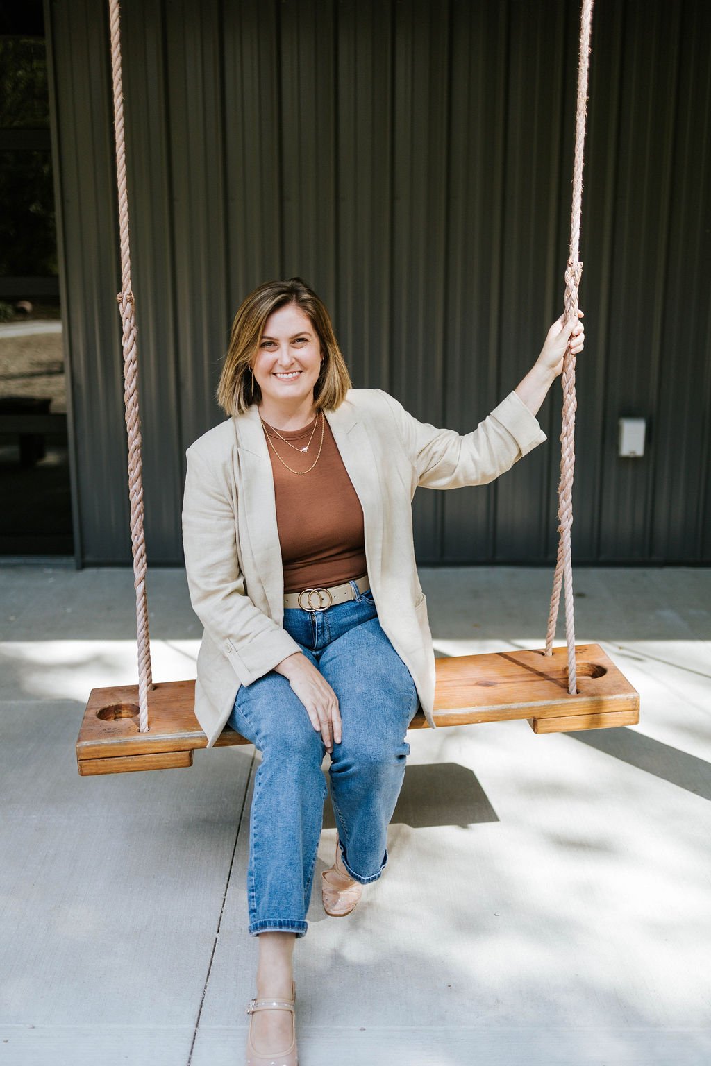 Jackie Burtch of Burtch Designs sitting on a wooden swing with ropes, outdoors against a black metal wall.