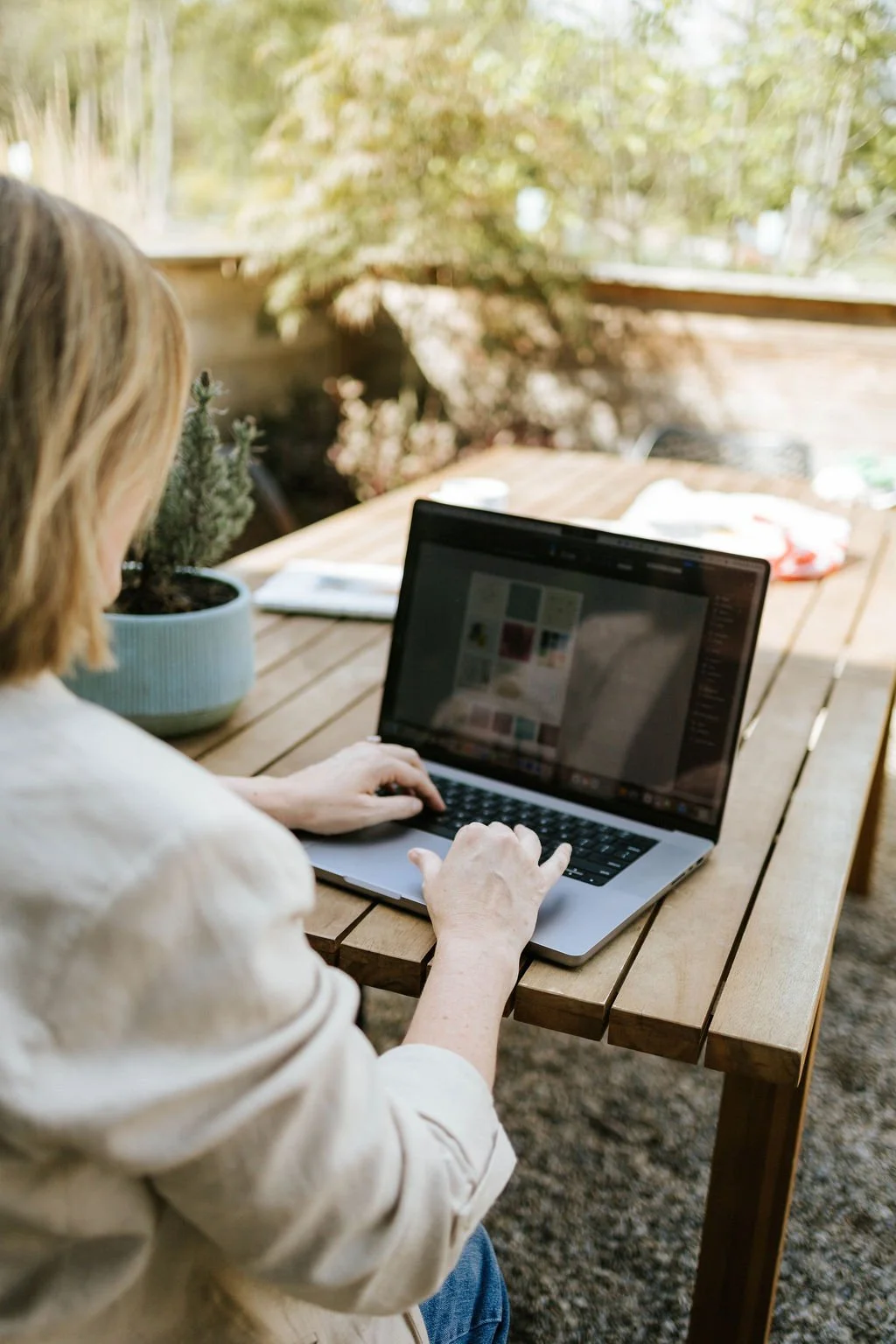 Jackie Burtch working on a laptop at an outdoor wooden table with a potted plant, surrounded by trees and natural light.