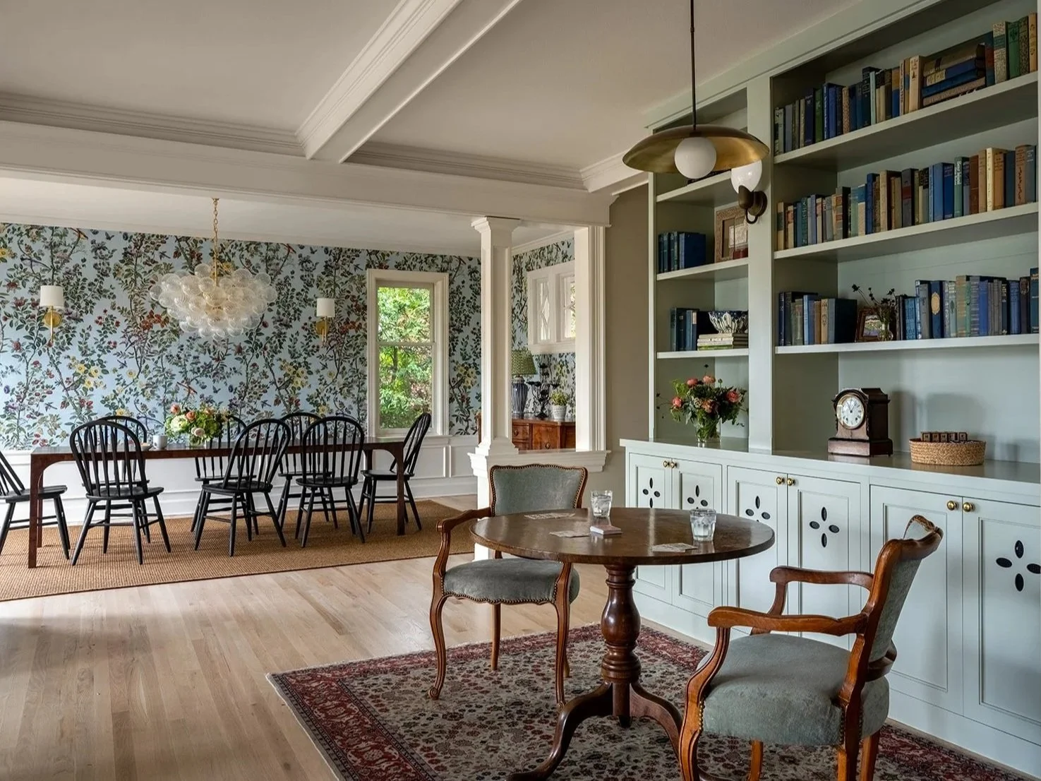 A spacious dining room with a long black wooden table, nine matching black chairs, floral wallpaper, light hardwood floors, a chandelier, and a built-in bookcase with books, decorative items, and flowers.