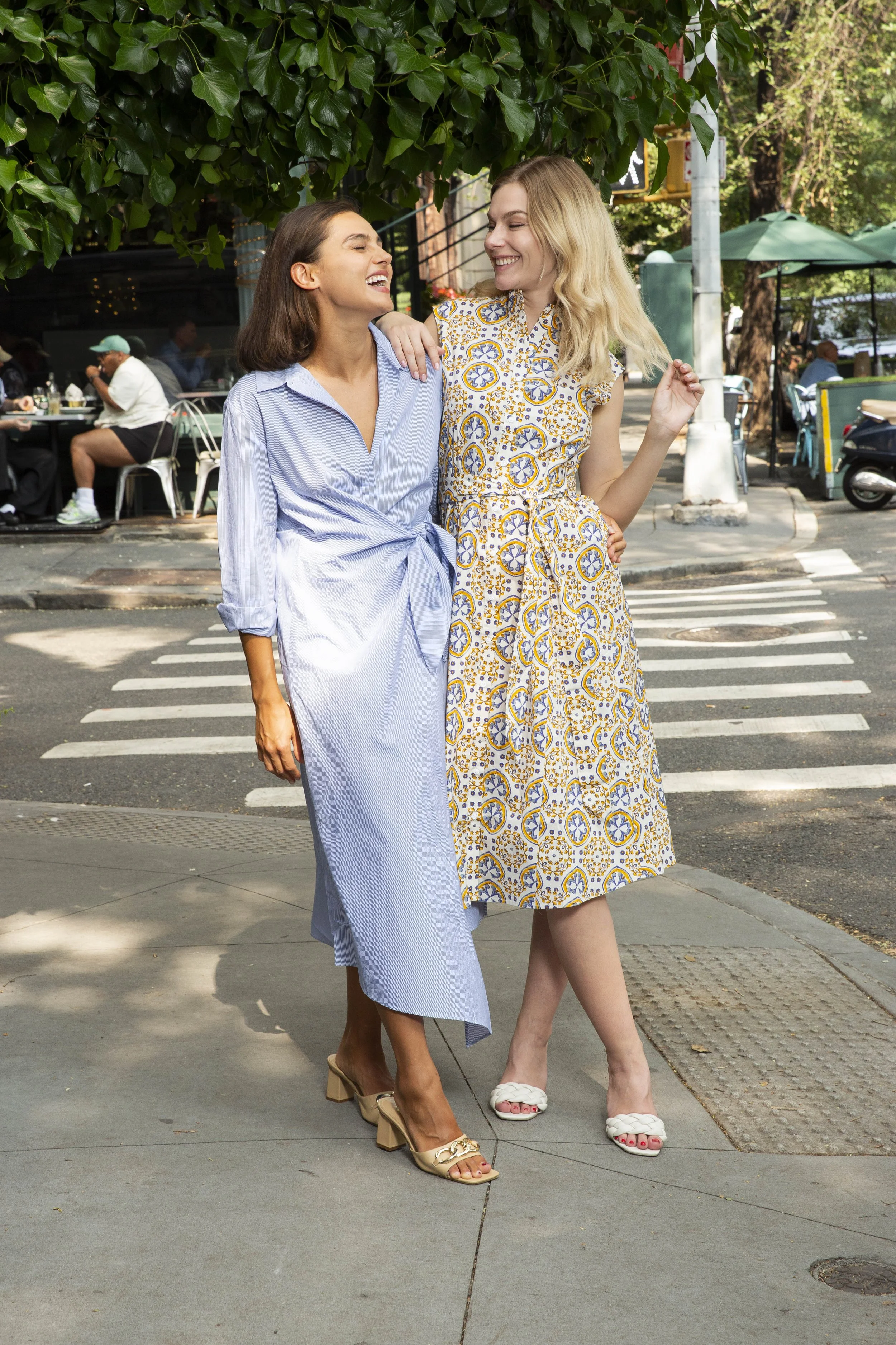 Two women smiling and walking arm-in-arm on a city street, dressed in summer clothing.