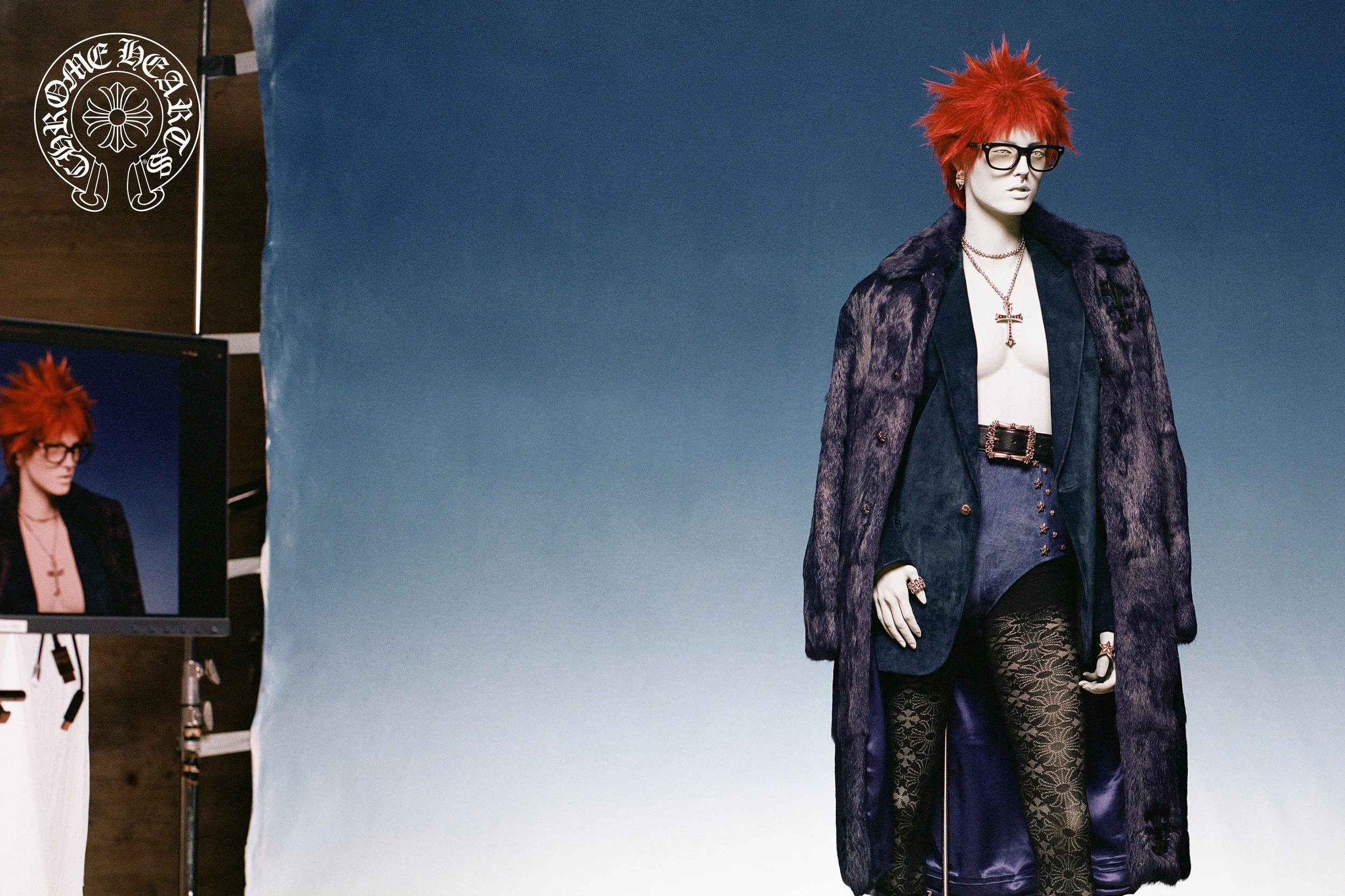 A mannequin with spiky red hair, black-rimmed glasses, and eclectic clothing, including a dark coat, patterned tights, and jewelry, against a gradient blue background.