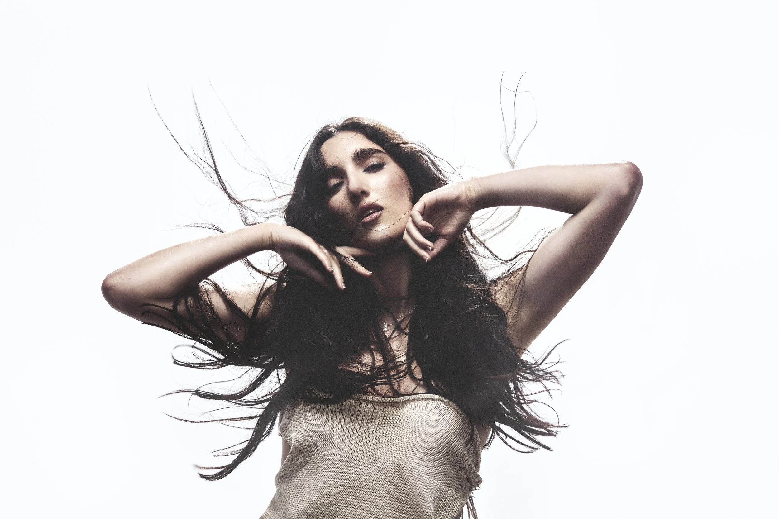 A woman with long black hair and light skin posing against a white background, with her eyes closed and her hands near her face.