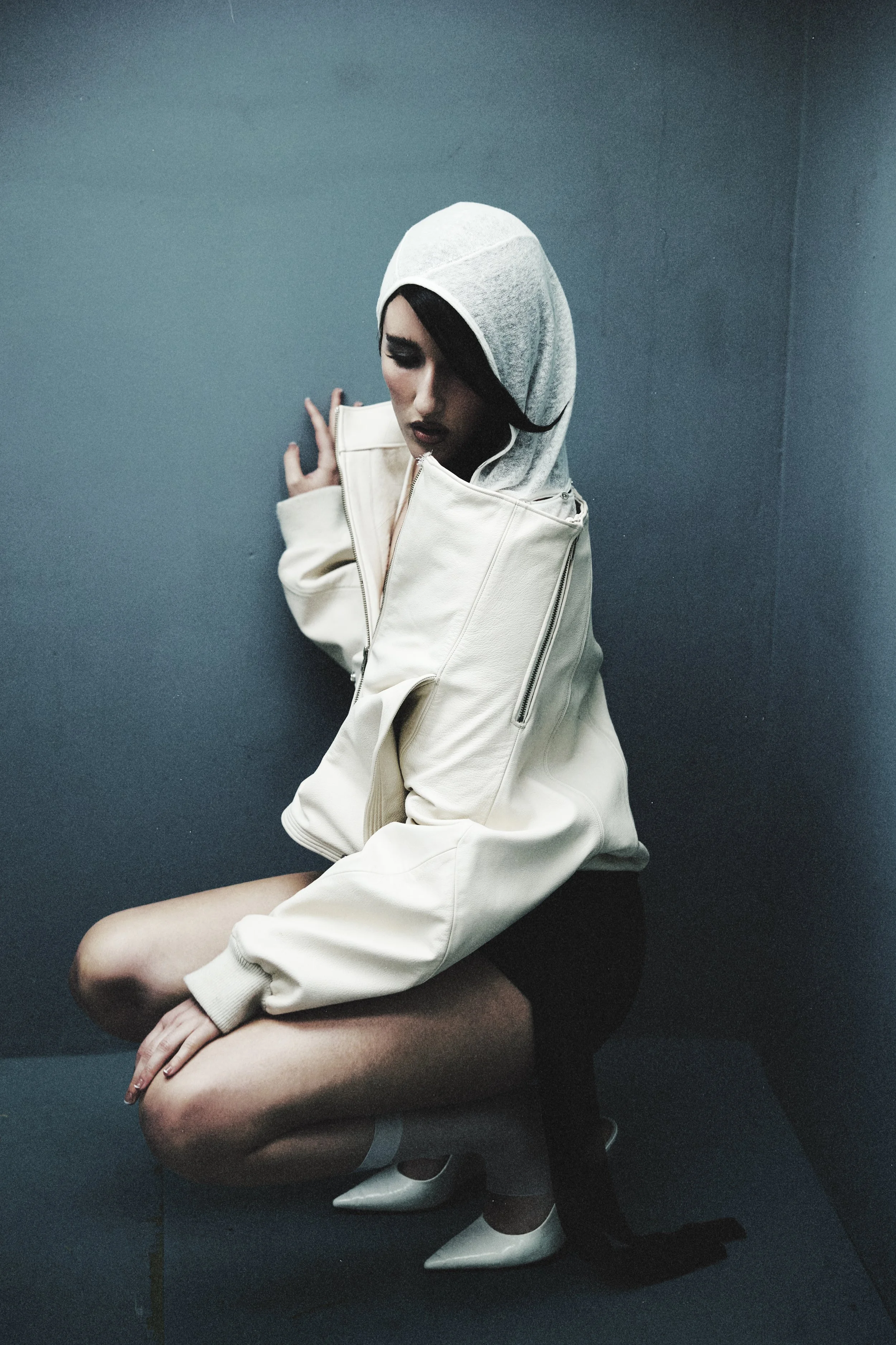 A woman crouches against a dark blue wall, wearing a white hoodie, white jacket, black shorts, white high heels, with dark makeup and short black hair.