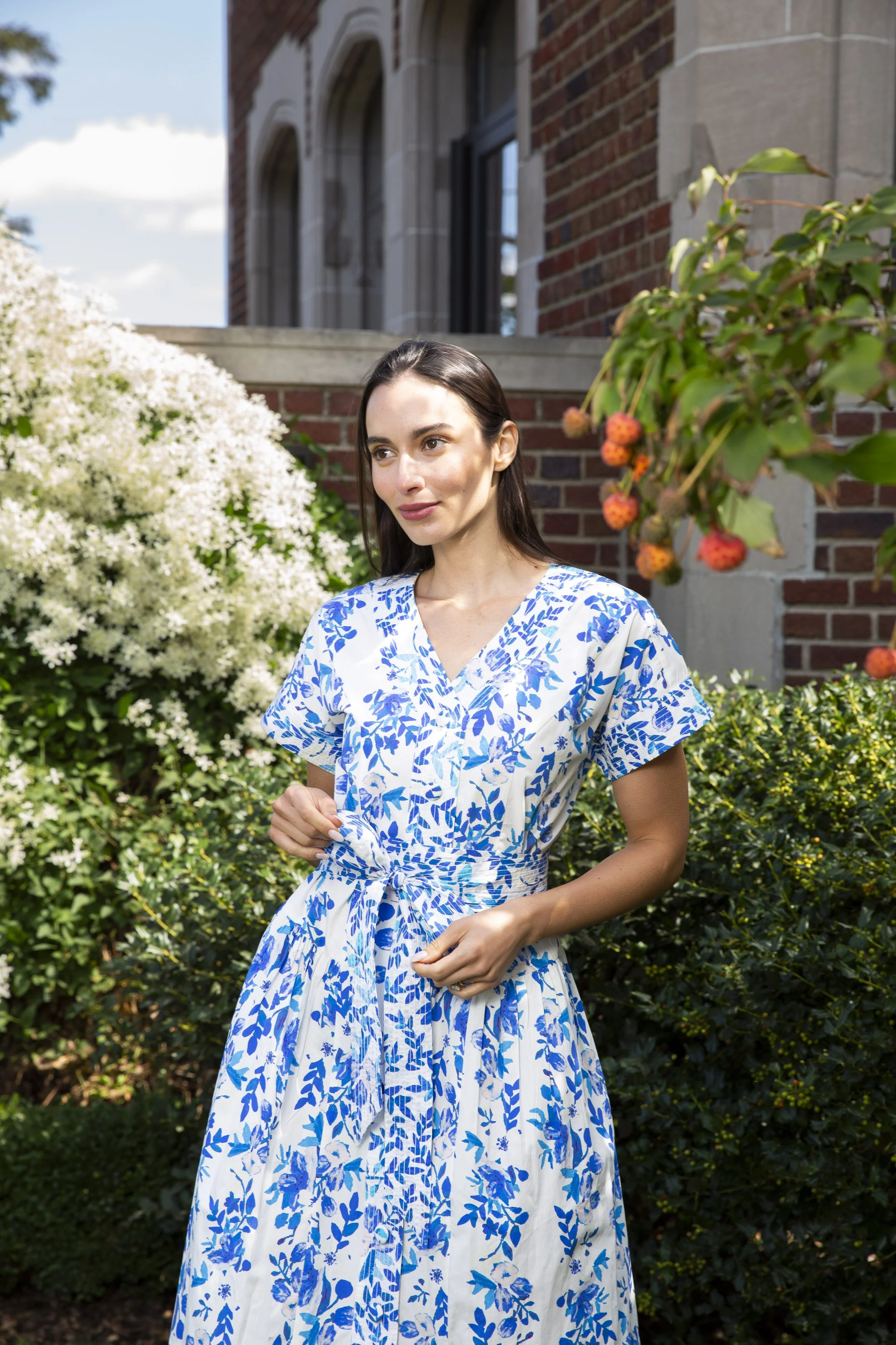 A woman in a white and blue patterned dress standing outdoors among green bushes and flowering plants, with a brick building in the background.