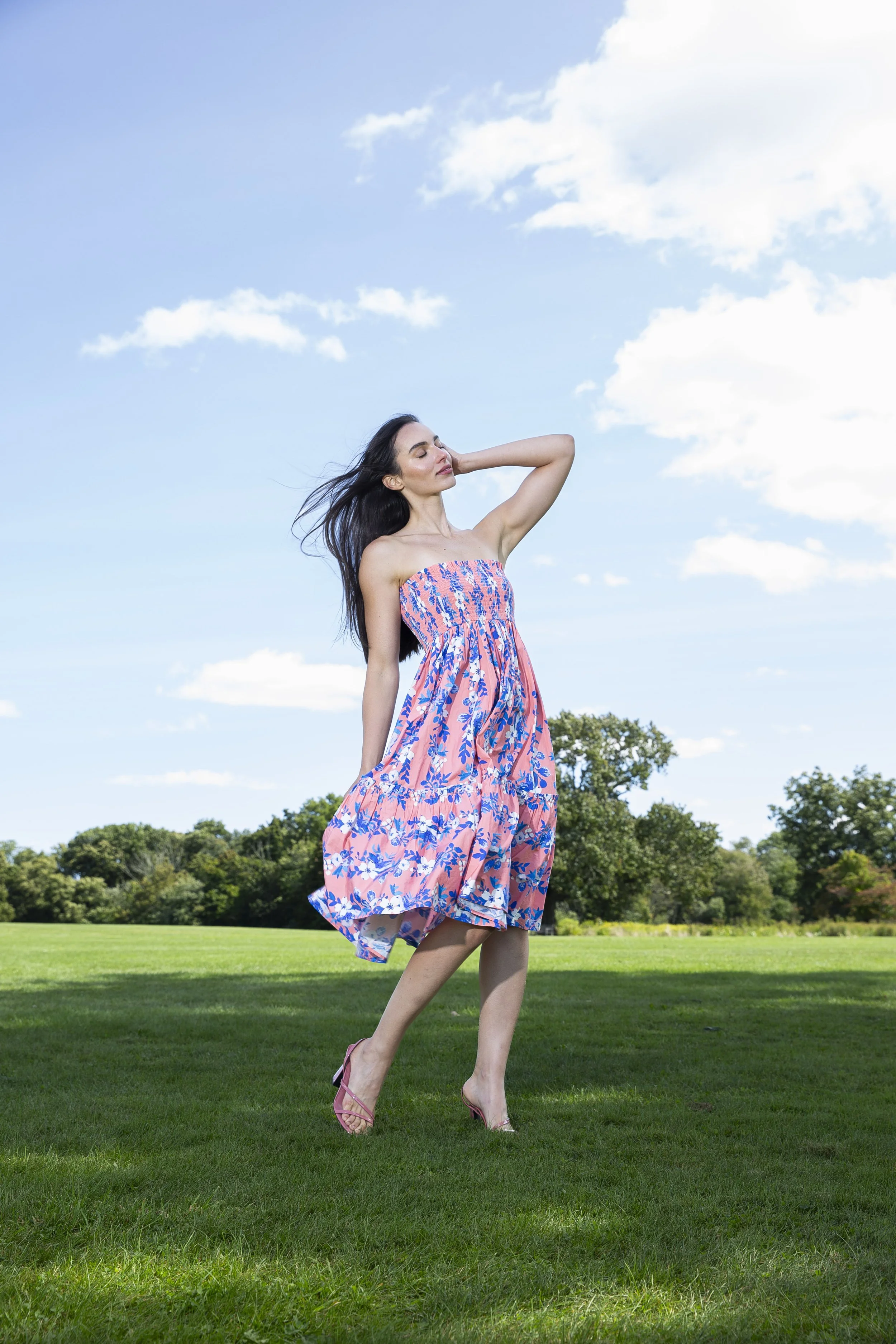Woman in a pink and blue floral dress standing on green grass under a blue sky with clouds, with trees in the background.