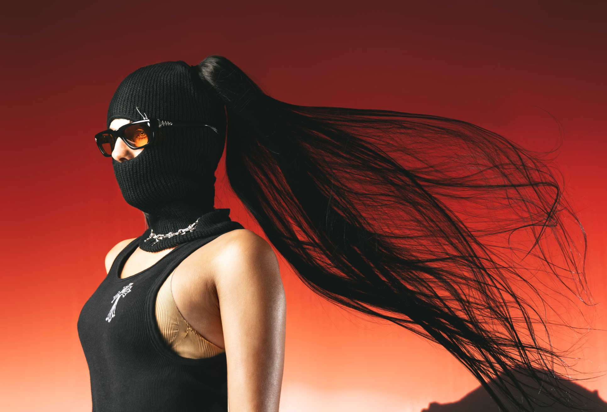 A woman with long black hair flowing in the wind, wearing sunglasses, a black face mask, a black sleeveless top with a small emblem, a chain necklace, and a black beanie hat against a red gradient background.