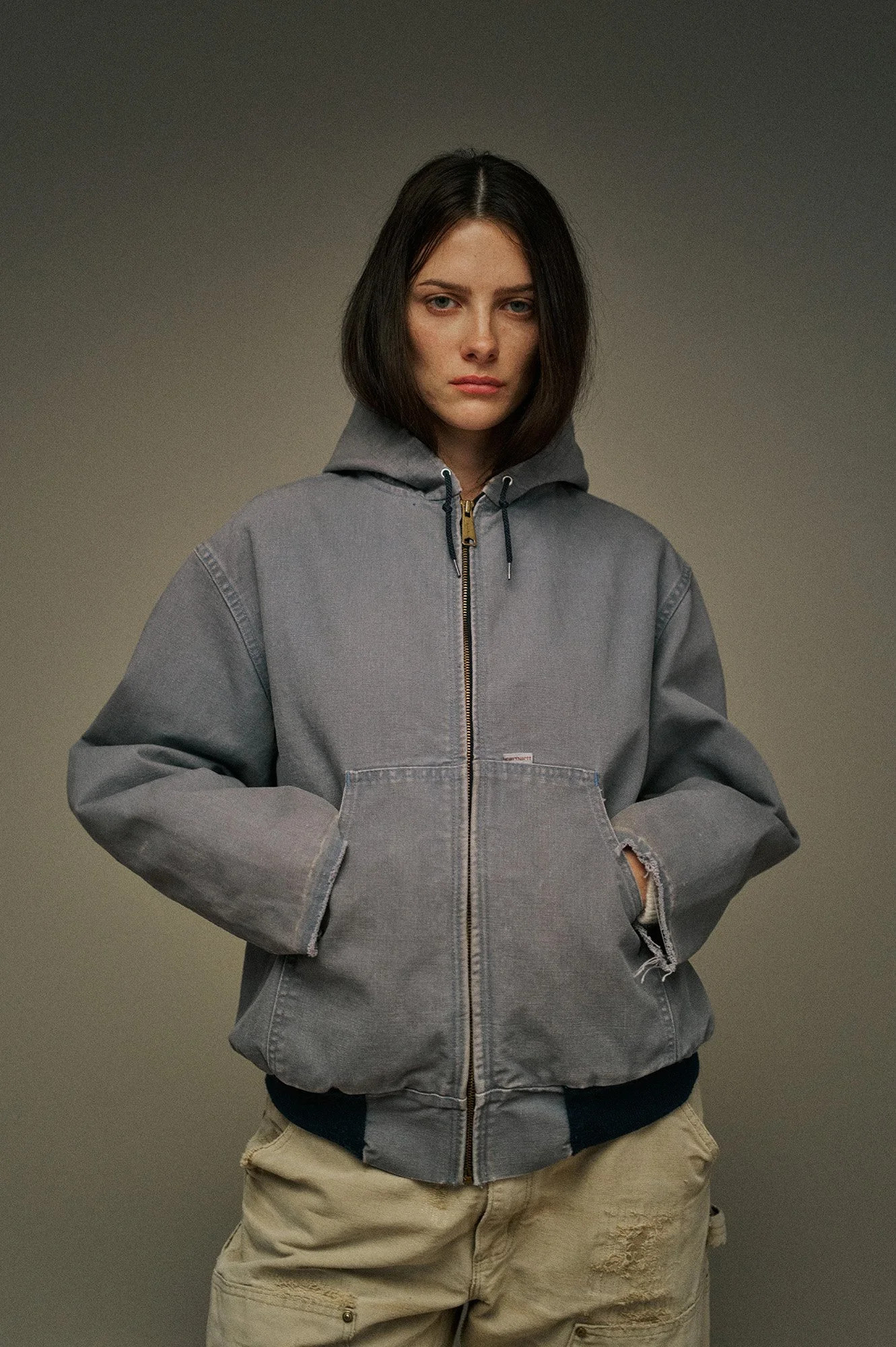 Studio fashion portrait by Scorpion Rose Studio & Lafayette American: Model in a distressed grey hooded work jacket and tan carpenter pants, highlighting high-end workwear aesthetic.