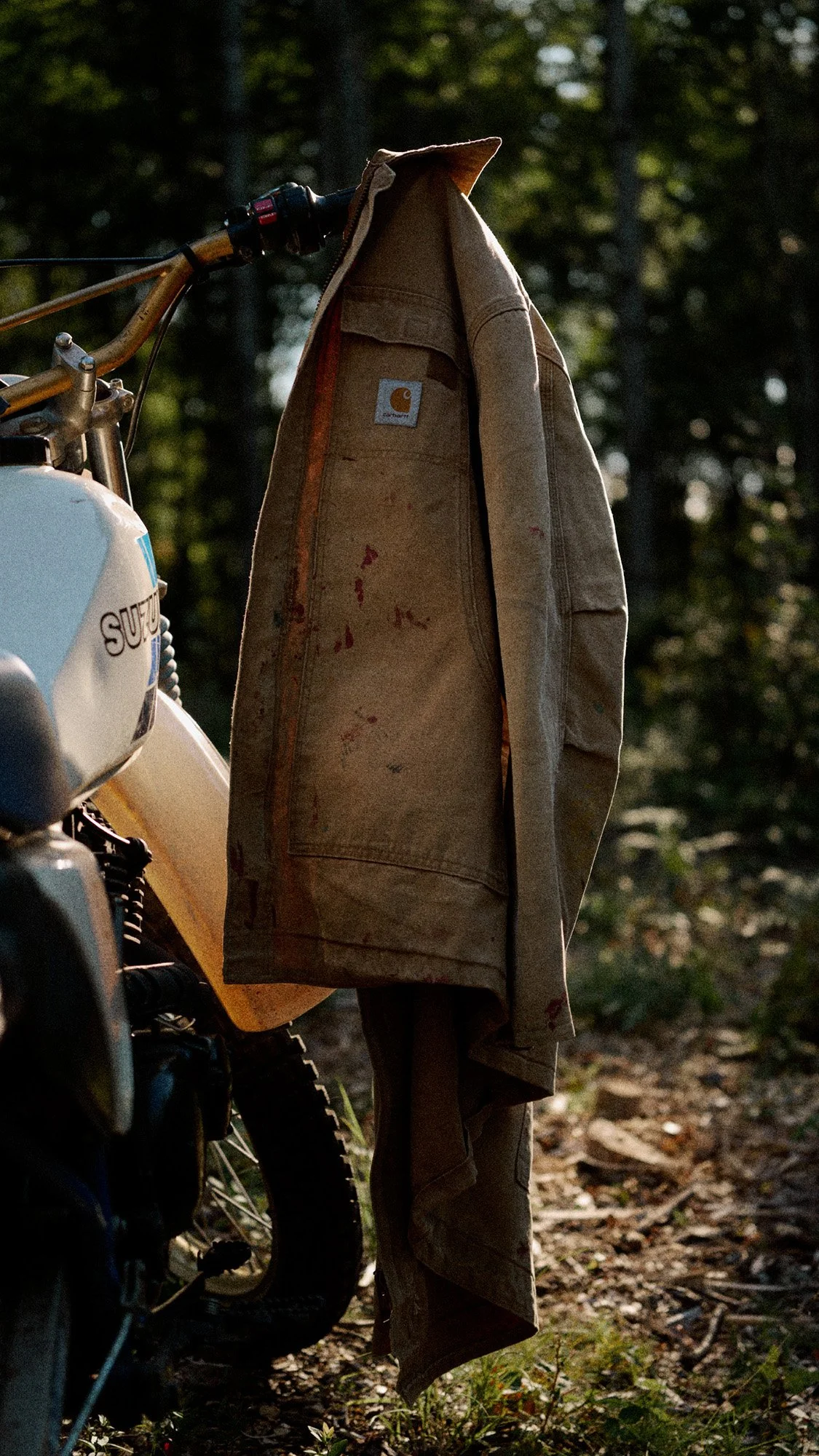 Cinematic lifestyle shot by Scorpion Rose Studio and Lafayette American: A distressed tan Carhartt jacket draped over a vintage Suzuki motorcycle in a sun-drenched wooded setting.