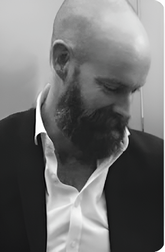 A bald man with a beard, dressed in a suit and white shirt, looking down.