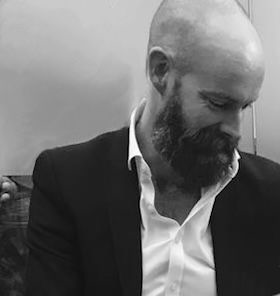 A man with a beard and buzz cut, wearing a dark blazer over a white shirt, looking downward, in a grayscale photo.