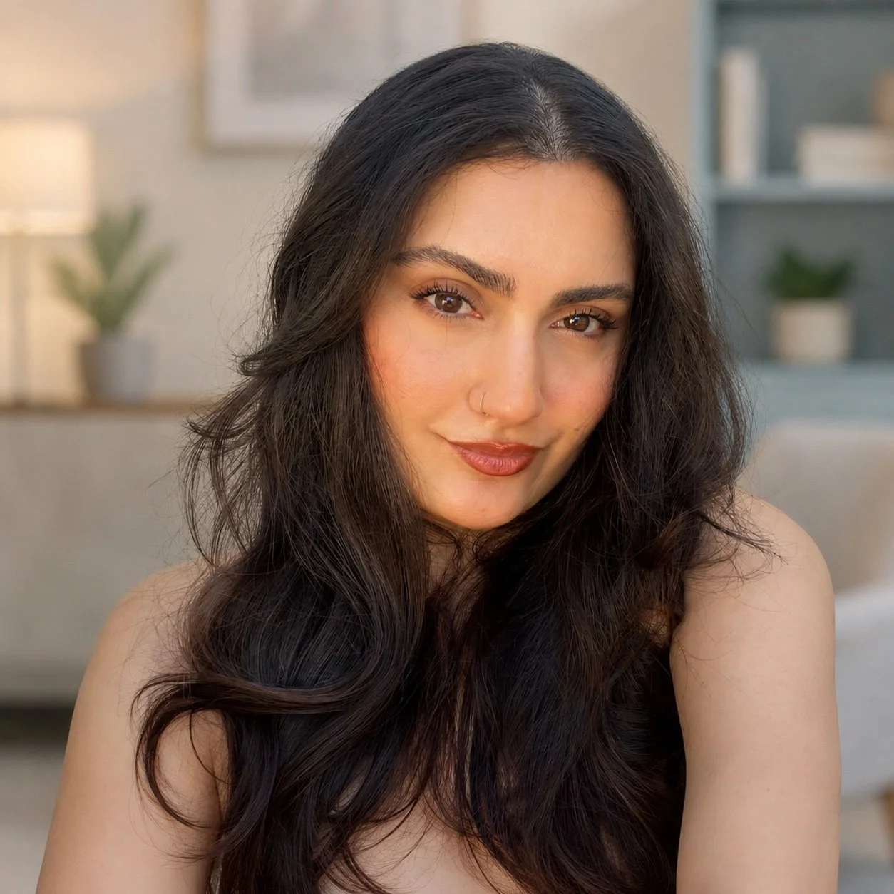 A young woman with long, dark wavy hair, warm brown eyes, and a subtle smile, posing in a cozy, well-lit room with a blurred background of shelves and a plant.