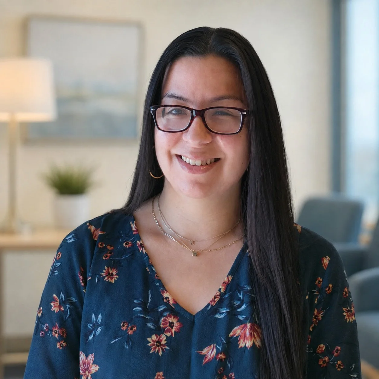 A woman with long black hair, glasses, and hoop earrings smiling in a well-lit room with a lamp, framed artwork, and a potted plant in the background.